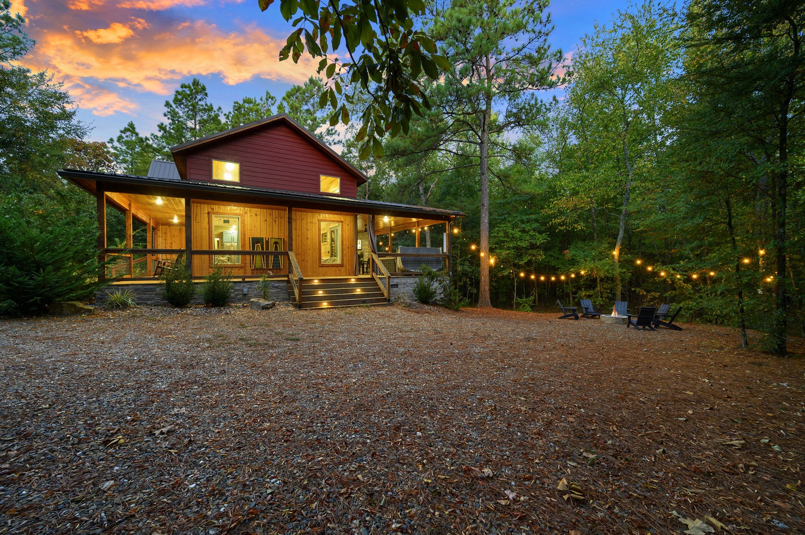 A charming cabin nestled in the woods welcomes guests at twilight, with string lights creating a warm ambiance across the outdoor gathering space and expansive parking area.