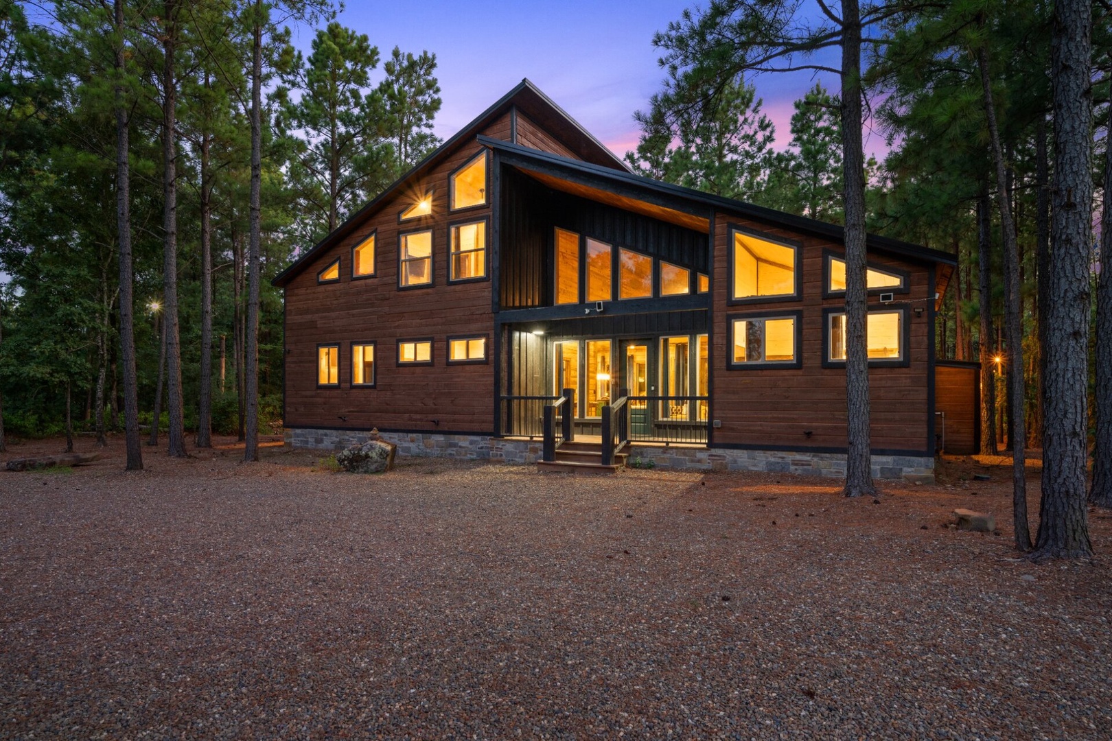 A modern wood-and-stone cabin nestled among towering pines, glowing warmly at dusk in a serene, forested setting.
