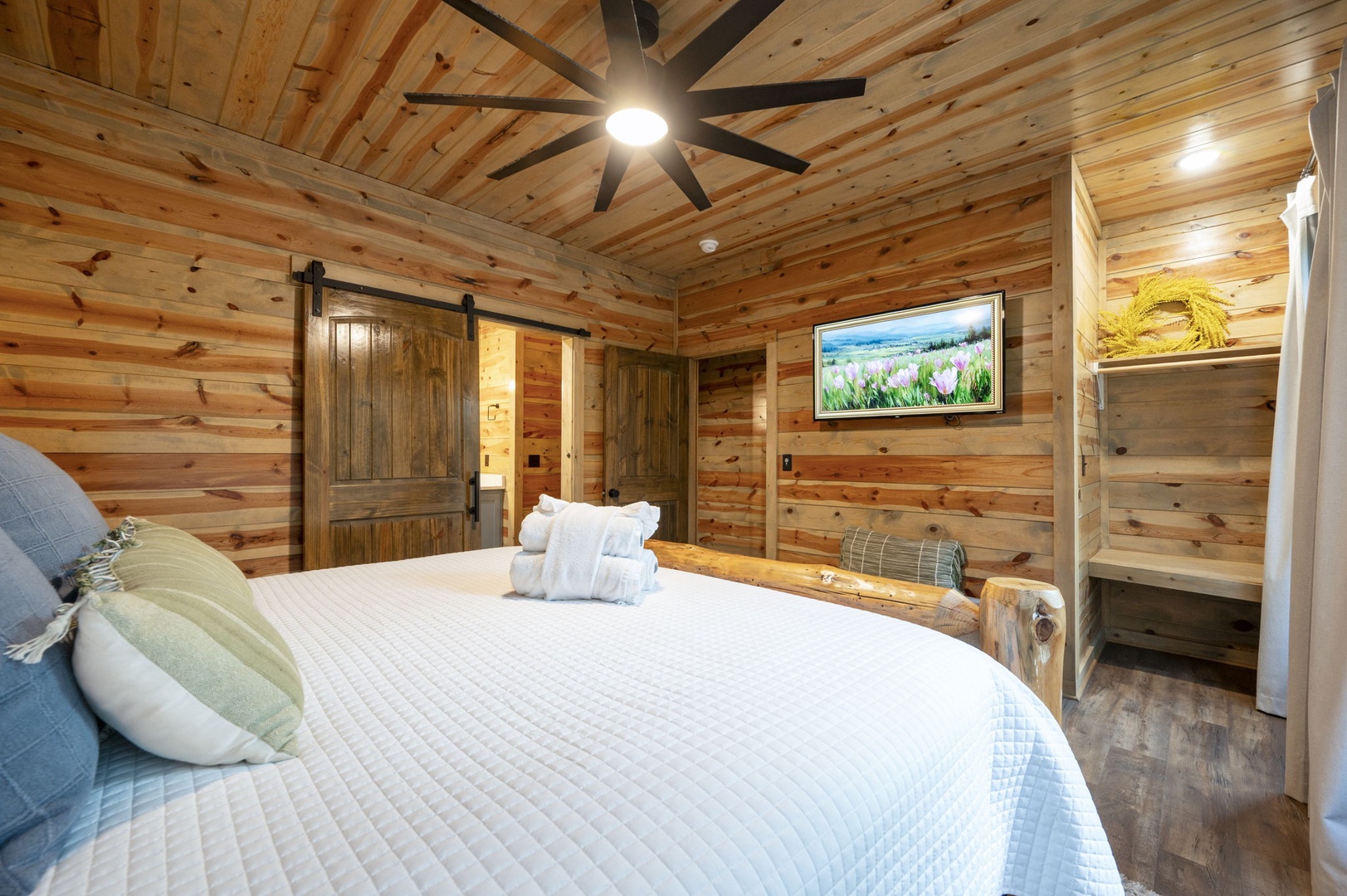 Warm wood surrounds you in this inviting bedroom retreat, where modern comfort meets rustic charm beneath a striking beamed ceiling and the gentle breeze of a ceiling fan.