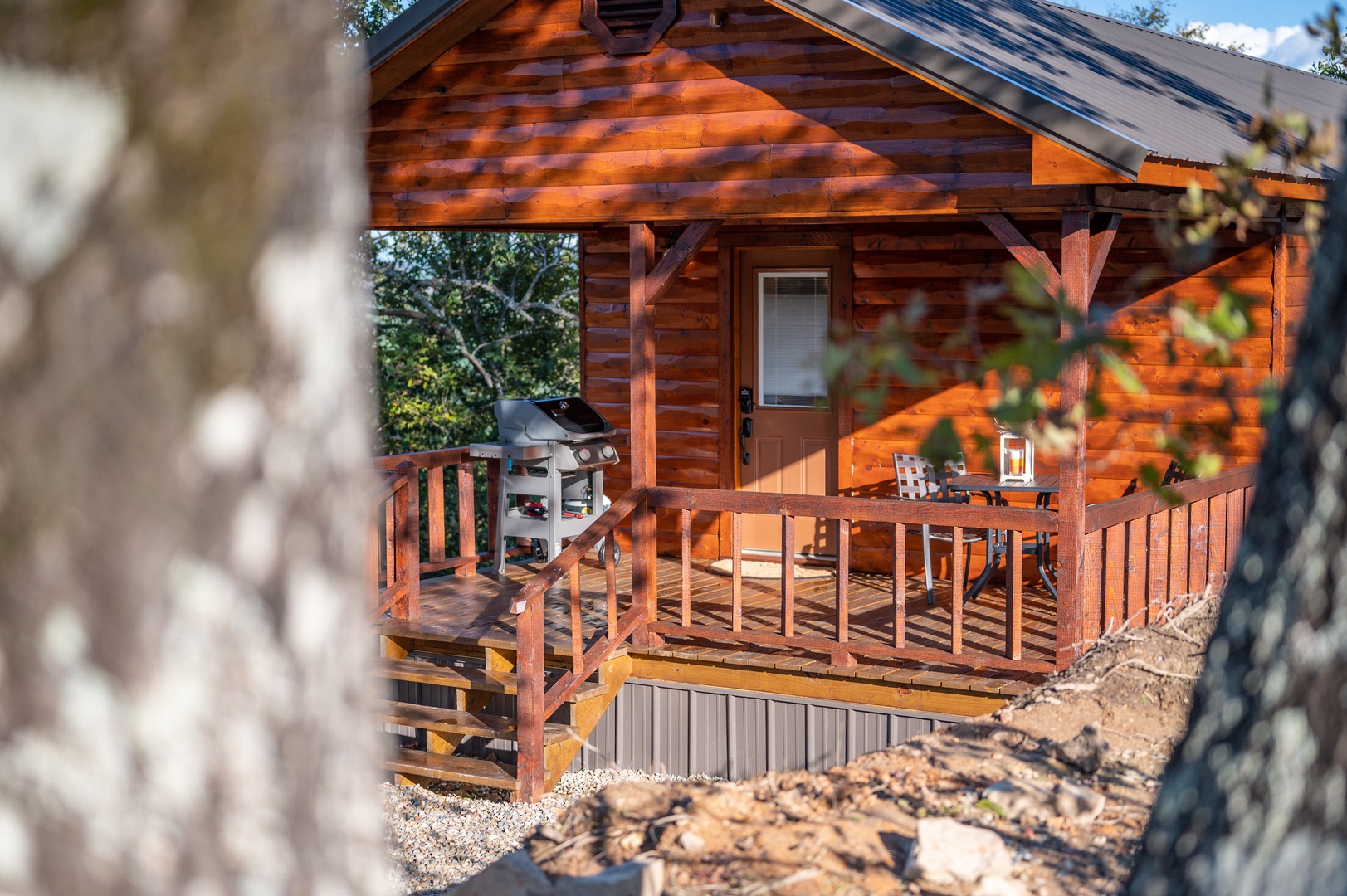 Rustic log cabin exterior featuring a spacious covered deck with BBQ grill and outdoor dining area, surrounded by natural landscaping.