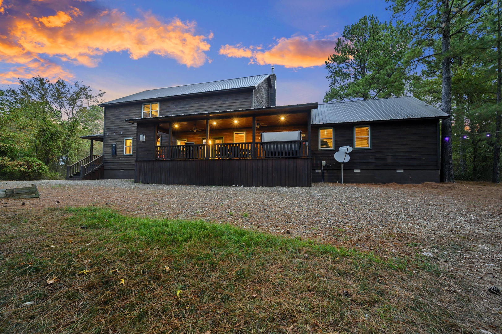 This charming cabin sits nestled among tall pines with a spacious gravel parking area, its warm interior lights glowing under a stunning sunset sky painted in vibrant orange and blue.