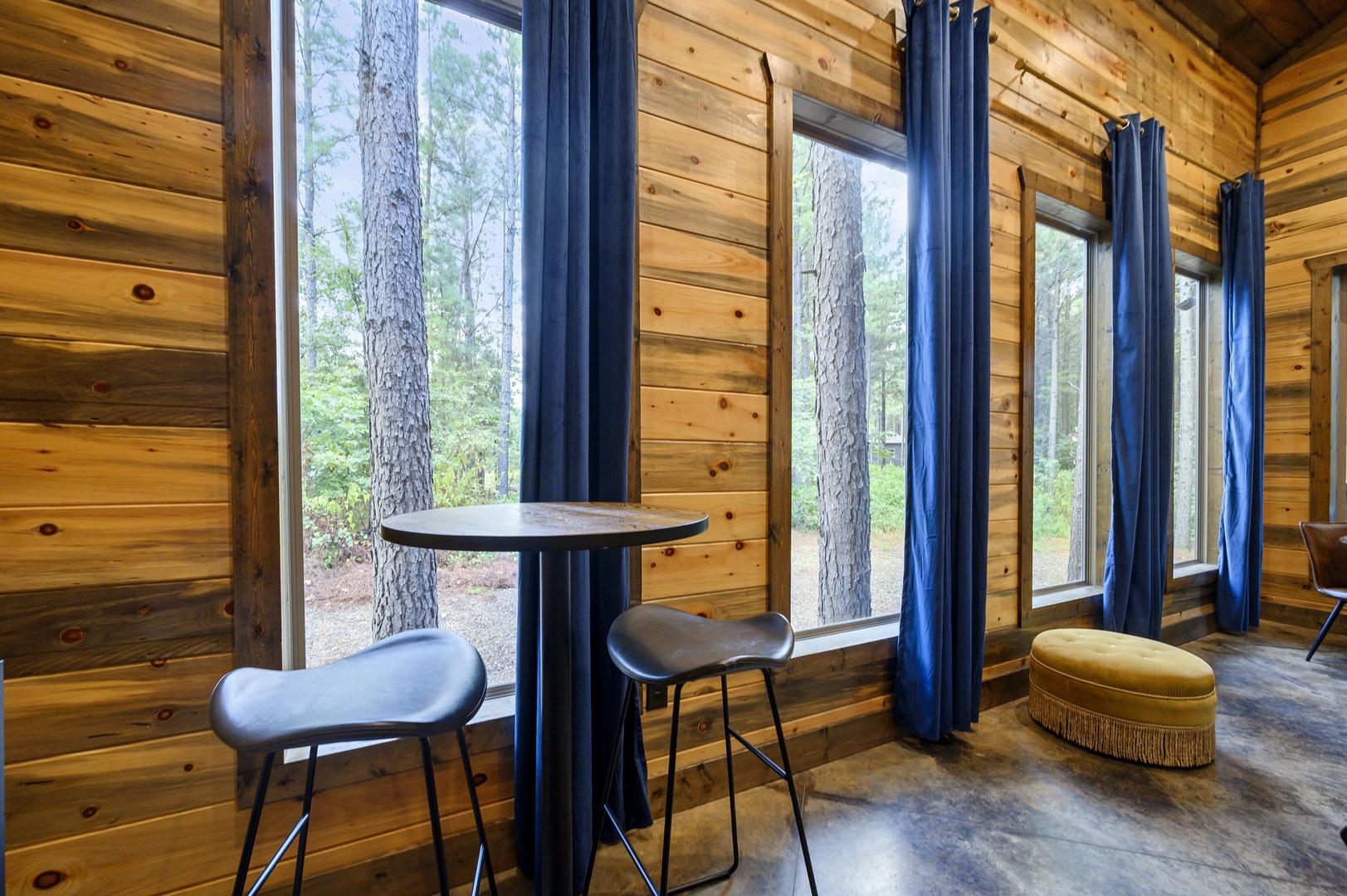 Step inside your tranquil forest retreat, where warm wood walls and expansive windows frame towering pines just beyond the glass. Cozy seating invites you to sip morning coffee while nature surrounds you.