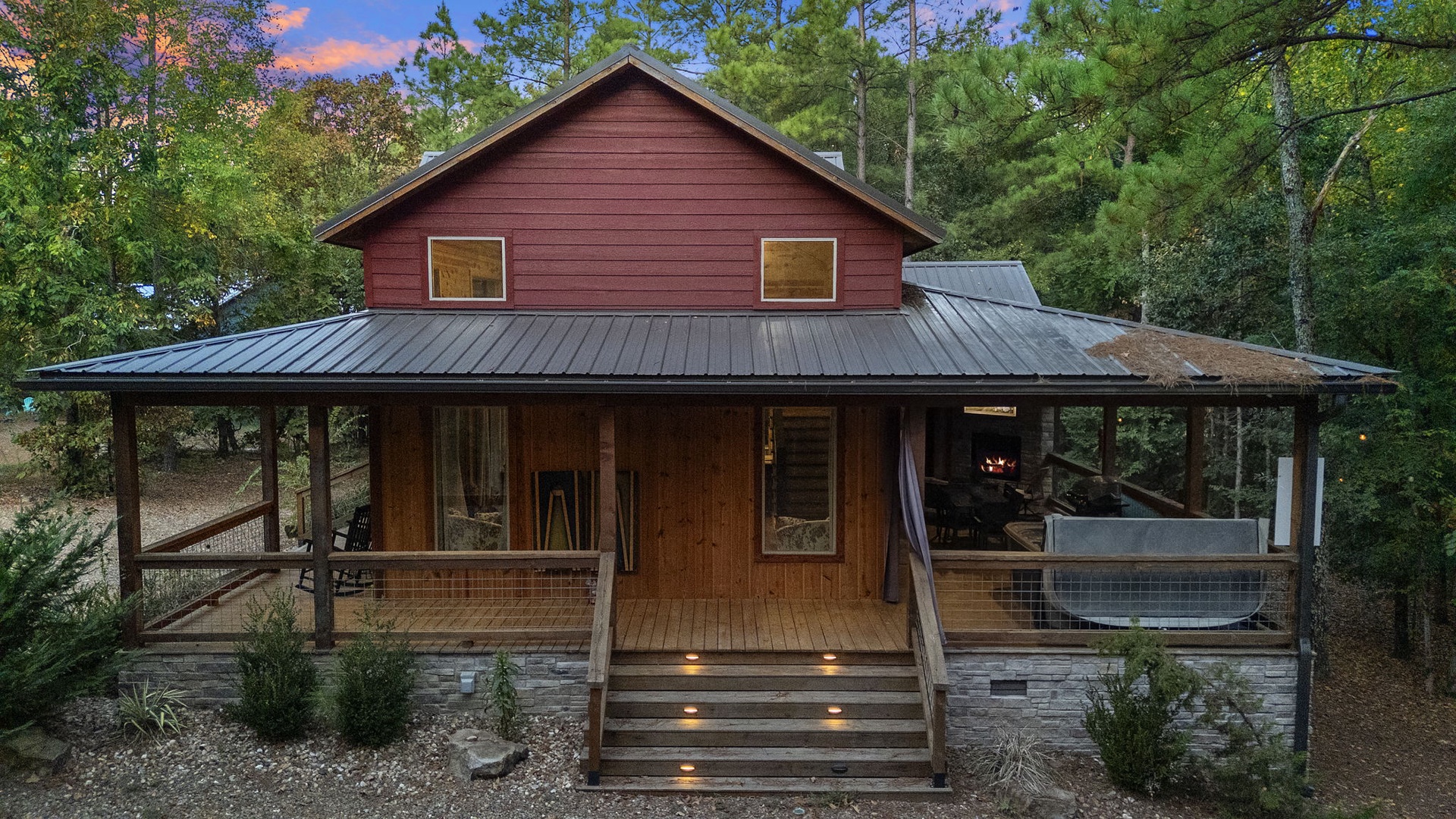 This charming red cabin is nestled among towering pines, offering a peaceful woodland retreat. The welcoming wraparound porch and soft evening sky create the perfect setting for your mountain escape.