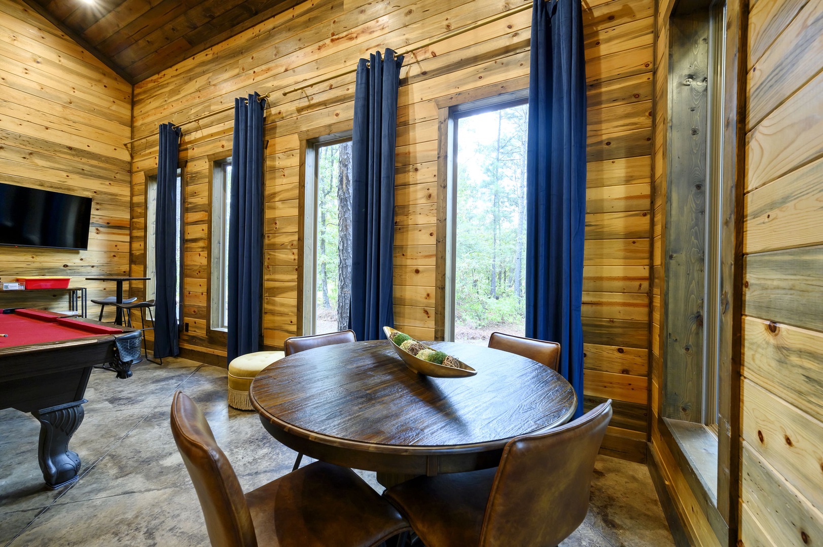 Gather around the handsome round table with forest views streaming through tall windows, while a pool table stands ready for your game nights in this warm wood-paneled retreat.