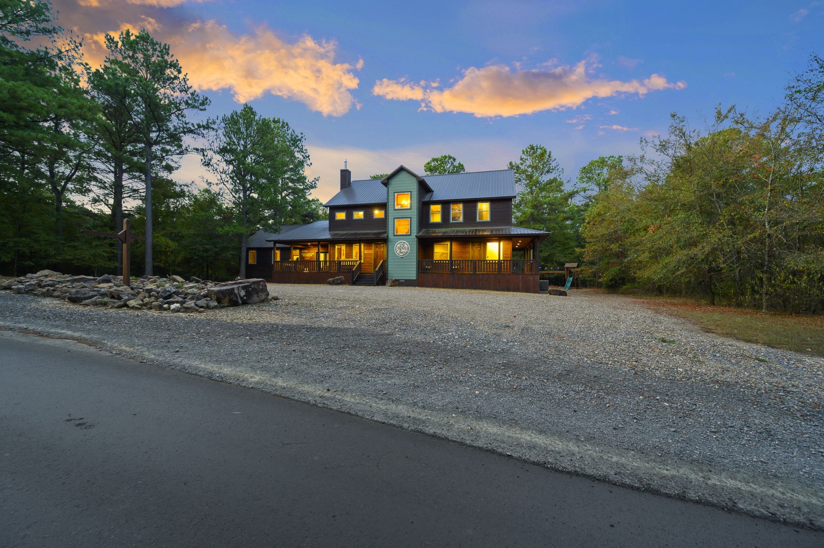 The property exterior glows warmly at dusk, surrounded by natural wooded scenery and ample gravel parking near the entrance.