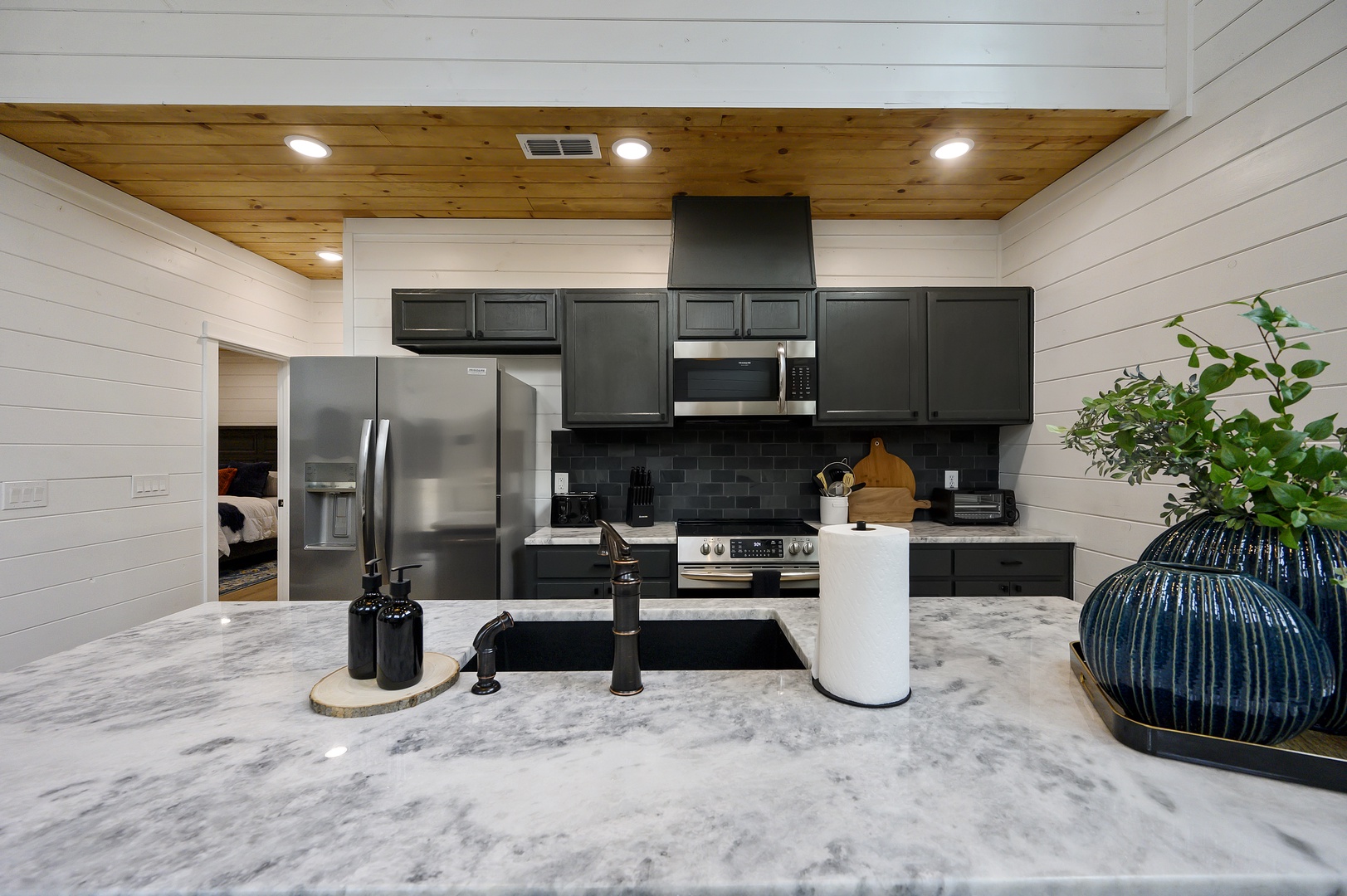 Cook with style in this modern kitchen featuring sleek dark cabinets, marble countertops, and stainless steel appliances beneath warm wood ceiling.