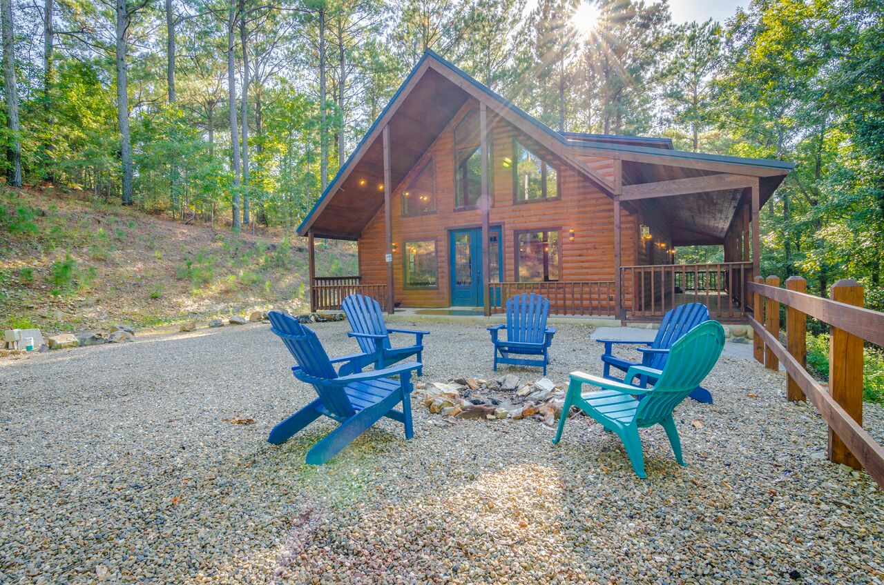 A modern cabin nestled in tall trees with a spacious gravel patio and colorful Adirondack chairs circling a stone fire pit, perfect for evening gatherings under the forest canopy.