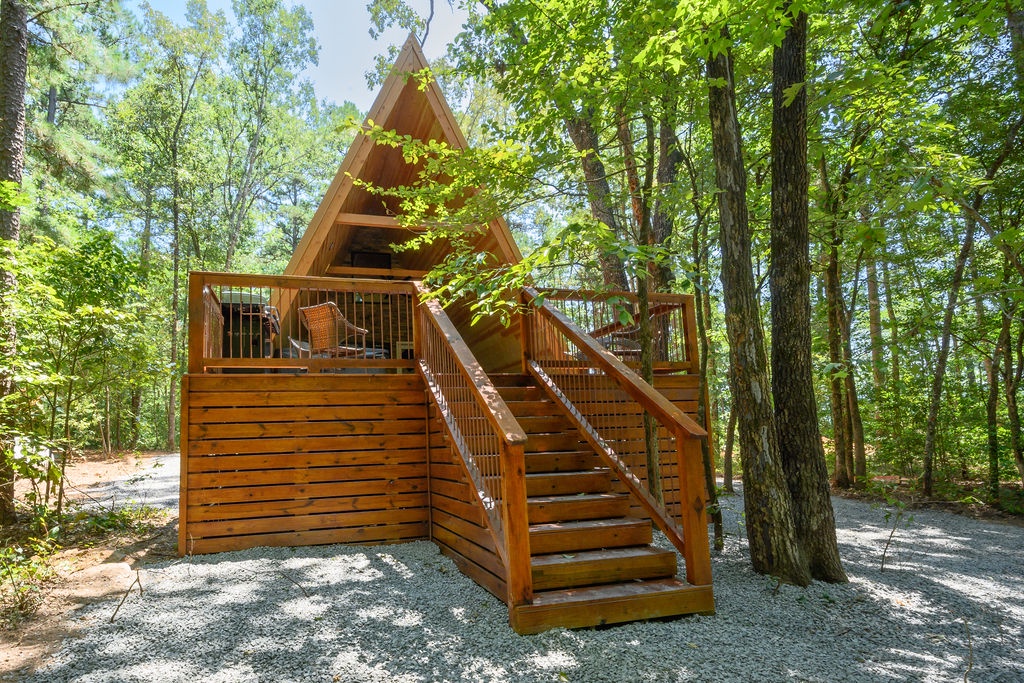 A charming A-frame cabin nestled among towering trees, featuring a private deck with seating where sunlight filters through the forest canopy. The natural setting offers peaceful seclusion and easy woodland access.