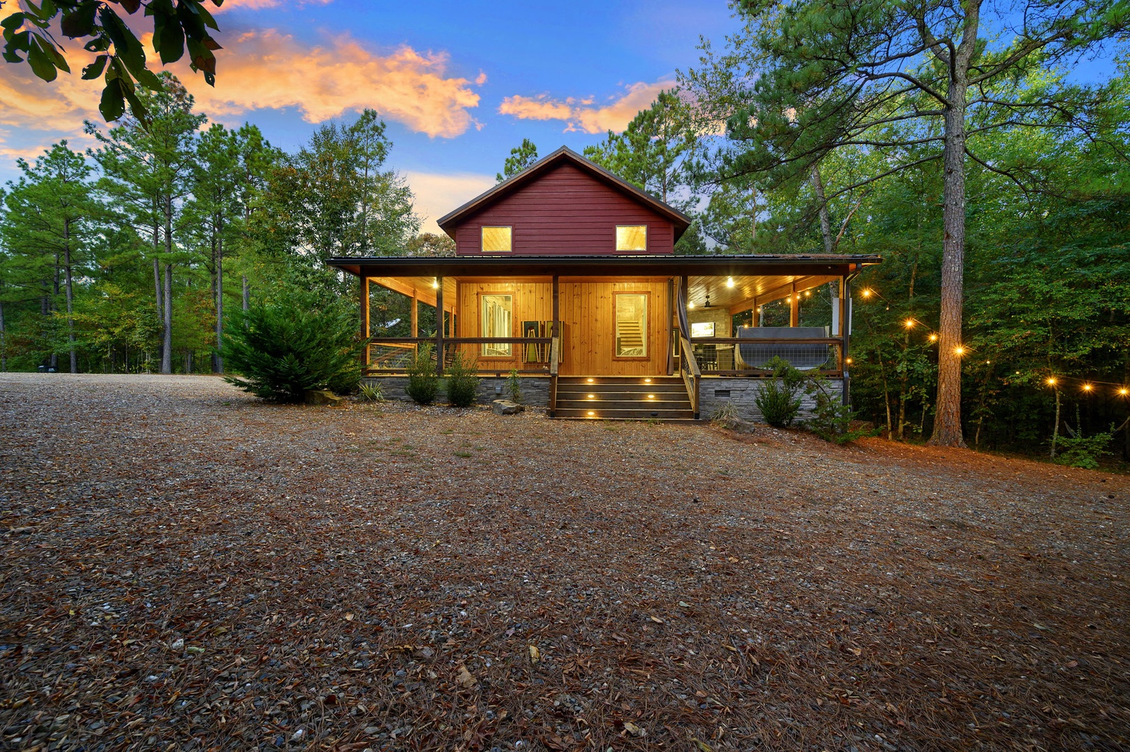 A charming cabin nestled among towering trees offers a warm retreat with inviting lights and a spacious covered porch, surrounded by peaceful woodland scenery at dusk.
