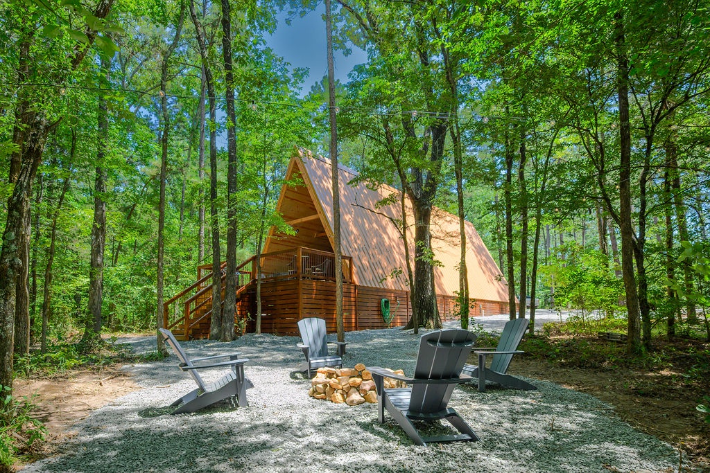 A cozy A-frame cabin nestled among towering trees, surrounded by a peaceful forest setting. The property features outdoor fire pit seating and a spacious deck for enjoying nature.