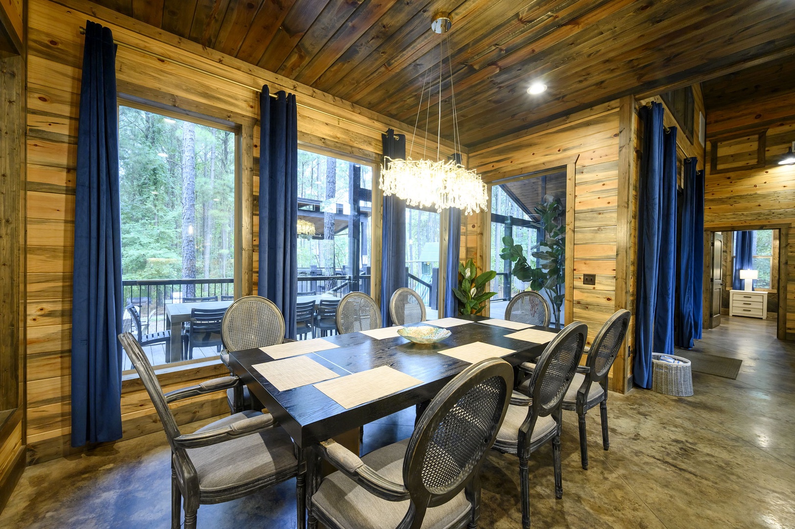 Step into your elegant dining room with warm wood paneling and a stunning chandelier overhead. Large windows frame peaceful forest views, while the spacious table invites memorable meals with loved ones.