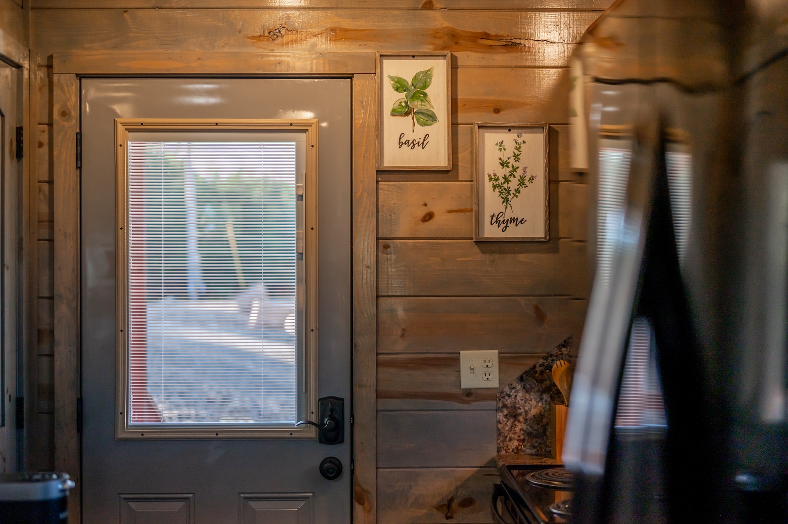 Warm up in your cozy kitchen featuring rustic wood walls, charming herb artwork, and natural light streaming through the door.