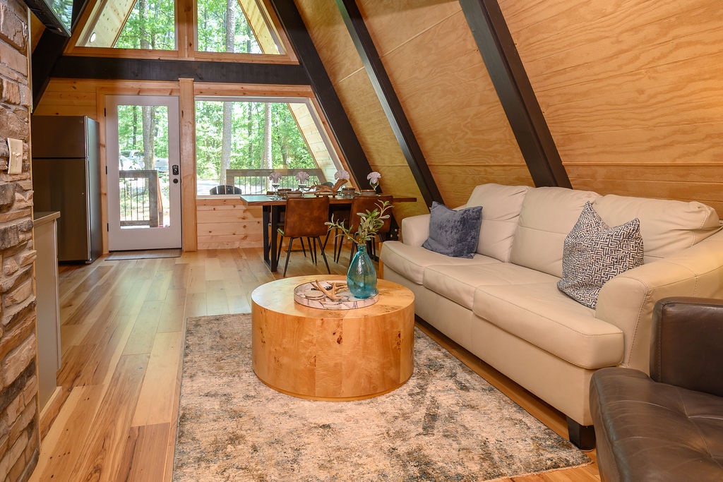 Step into your cozy A-frame retreat where soaring ceilings and floor-to-ceiling windows frame peaceful forest views. Gather around the dining table or sink into the plush seating—this sunlit space invites you to unwind in natural beauty.