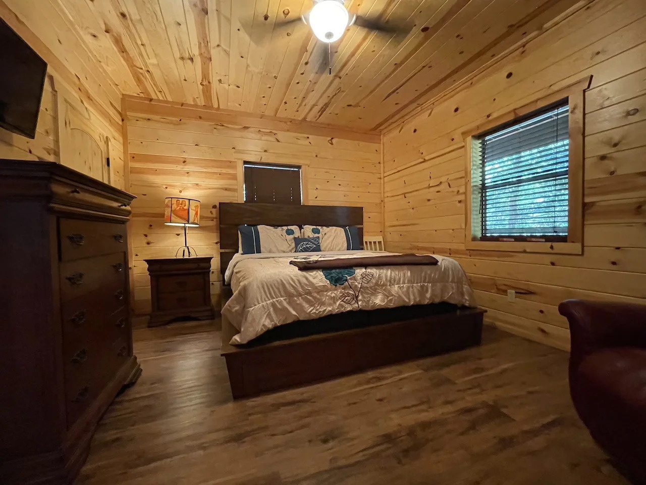 Your cozy cabin bedroom awaits with warm pine walls, a plush bed dressed in soft linens, and gentle natural light filtering through the window—perfect for peaceful mountain mornings.