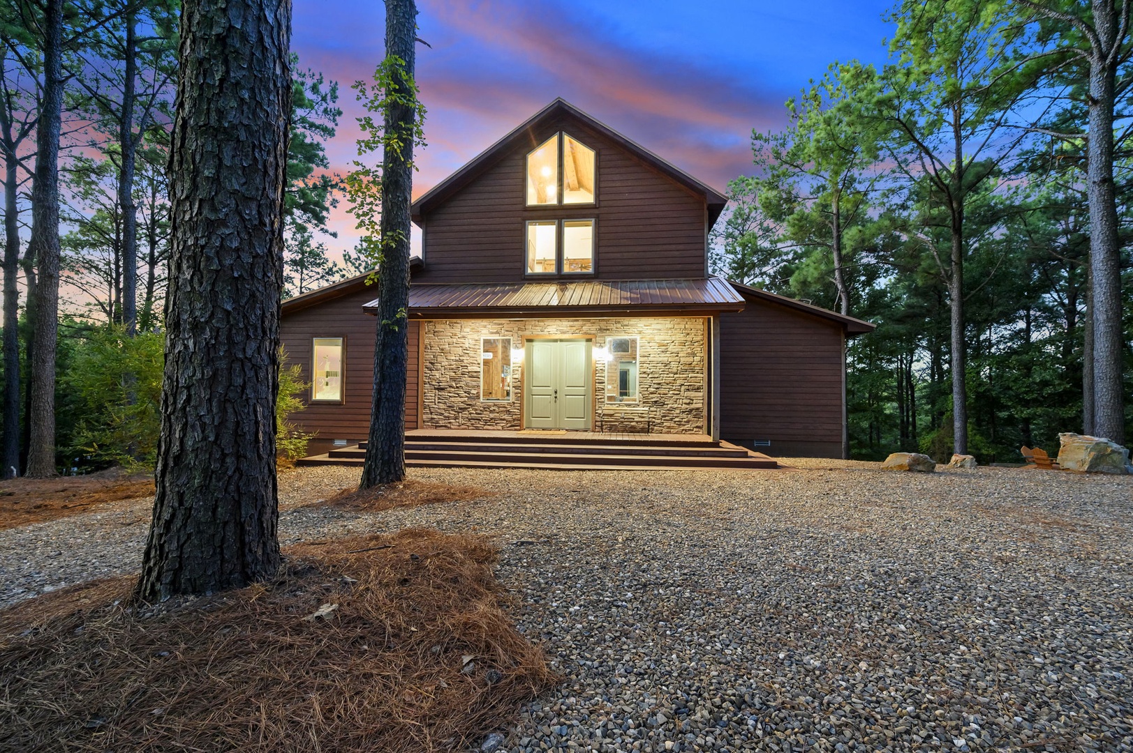 A striking modern cabin nestled among towering pines, featuring warm stone accents and a welcoming entrance. The property combines rustic charm with contemporary design, set against a peaceful wooded backdrop at dusk.