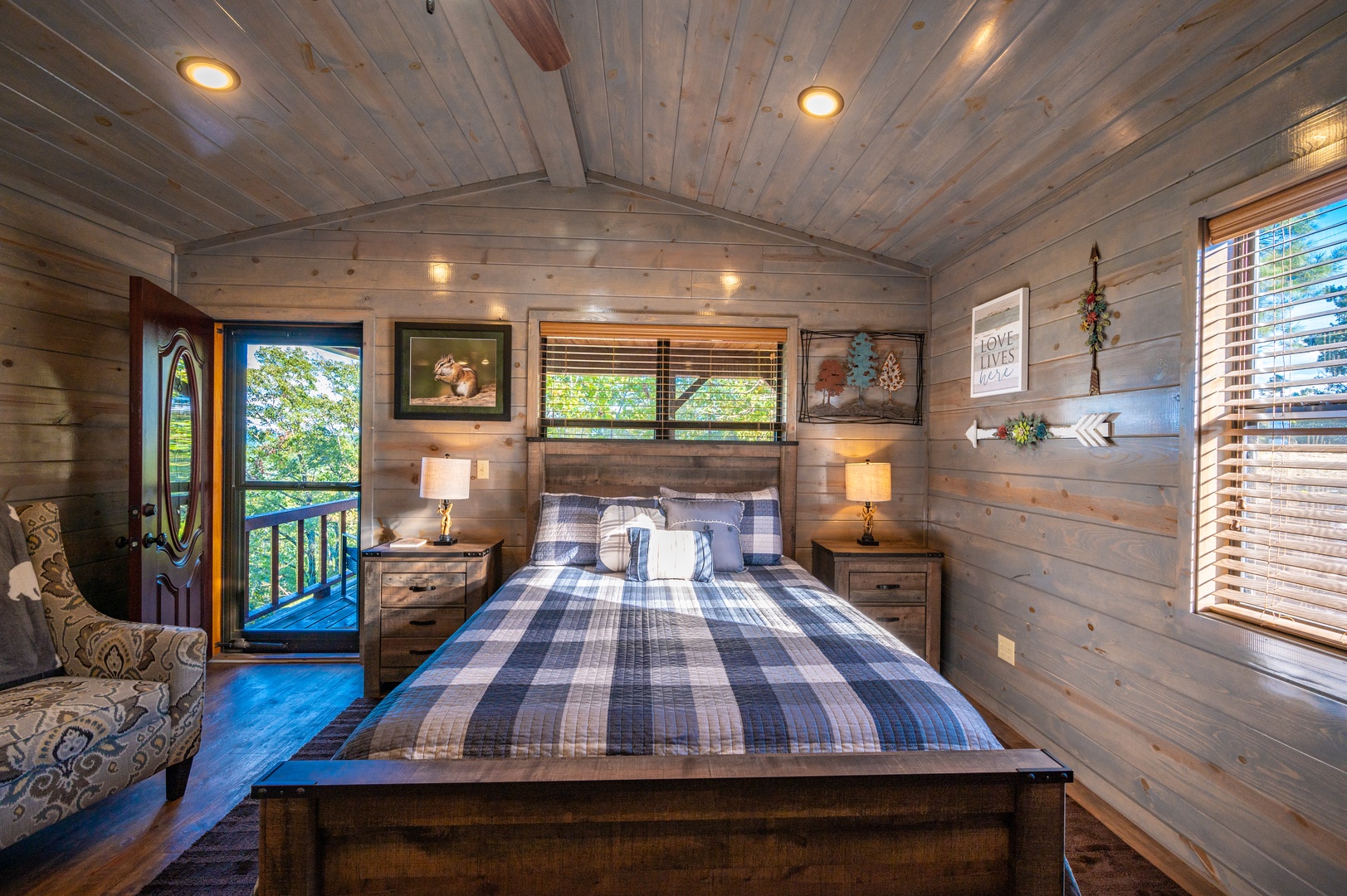 Relax in your cozy cabin bedroom with rustic wood walls, plaid bedding, and private balcony overlooking lush greenery.
