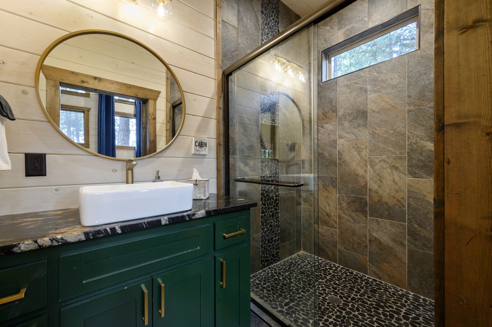 Spa-like sanctuary with warm wood tones, elegant stone tile, and gold accents. Walk-in shower with riverstone flooring and vessel sink on green vanity beneath a striking round mirror.