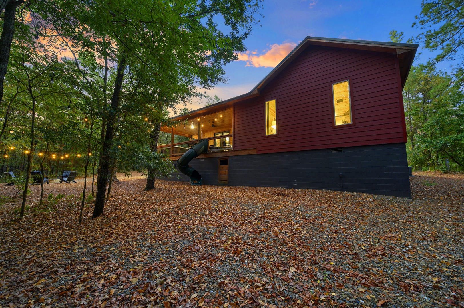 A modern cabin tucked among towering trees, glowing warmly as dusk settles over the surrounding woods and autumn leaves blanket the forest floor.