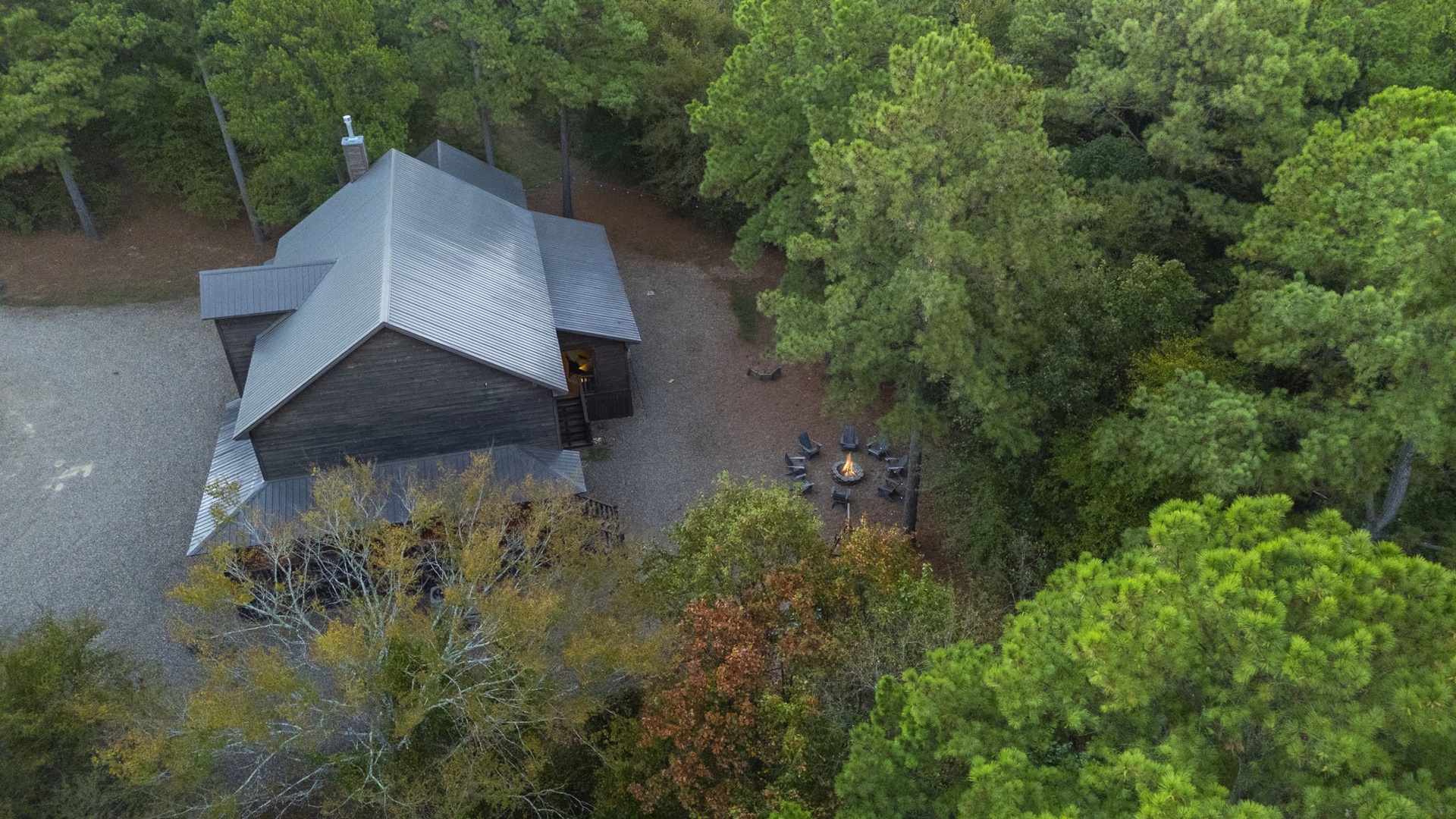 Aerial view of a secluded modern cabin nestled among lush forest, with a cozy outdoor fire pit area perfect for peaceful evenings in nature.