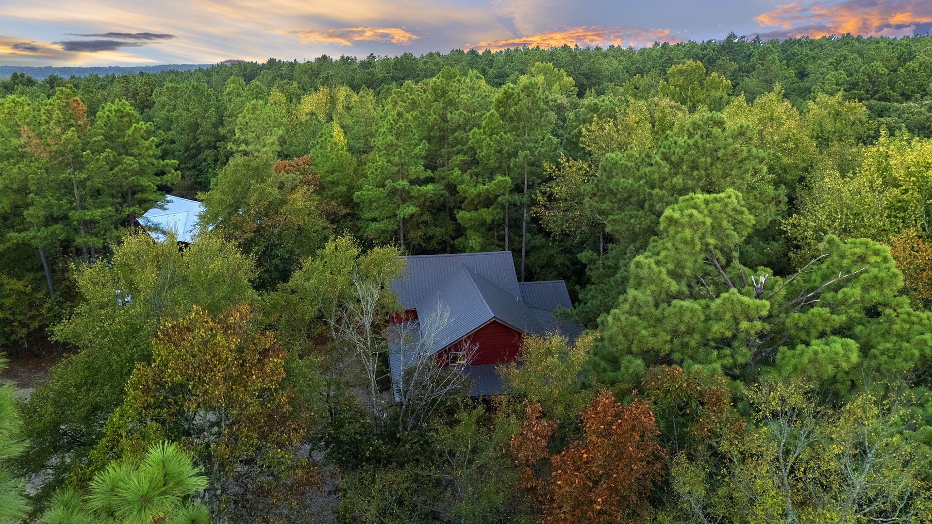 Aerial view of a charming red barn nestled in a lush forested setting beneath colorful sunset skies, showcasing the natural beauty surrounding the property.