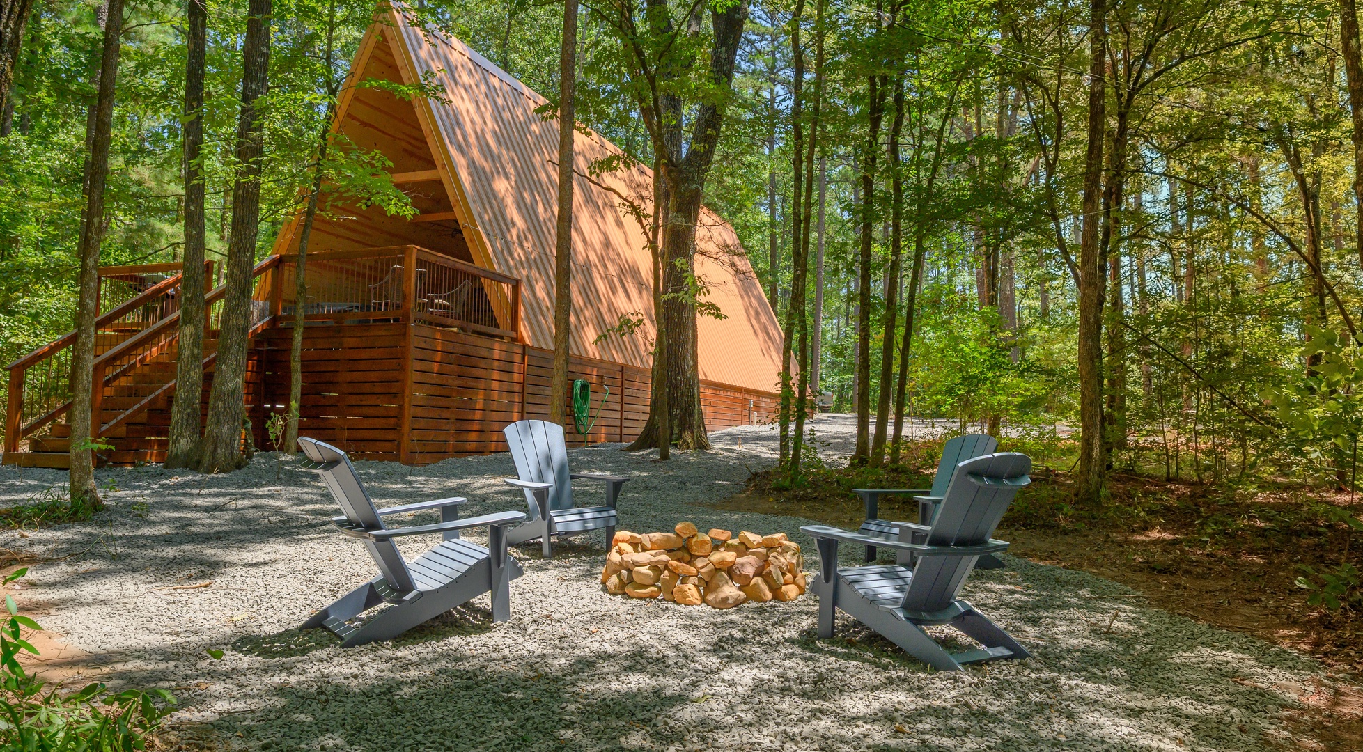 A cozy A-frame cabin nestled among towering trees, surrounded by a peaceful forest setting. The property features outdoor fire pit seating and a spacious deck for enjoying nature.