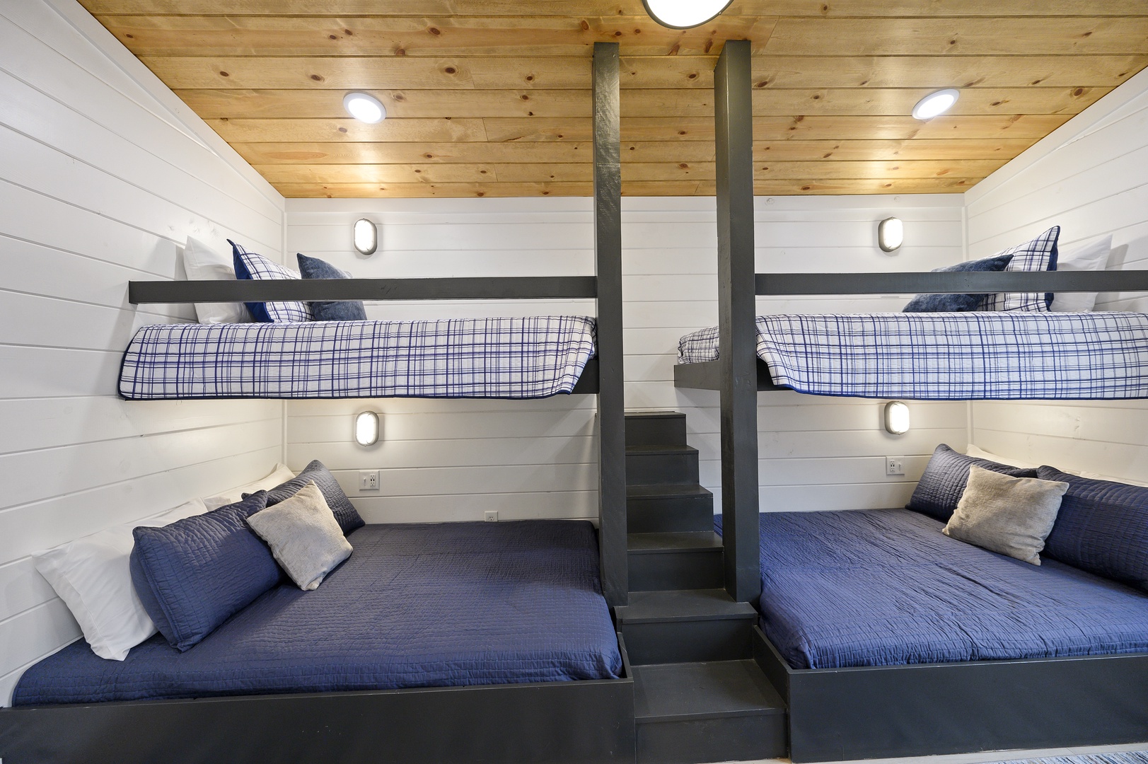 Your cozy bunk room offers comfortable sleeping for four, with custom-built beds featuring reading lights and plaid accents beneath warm wood ceilings.