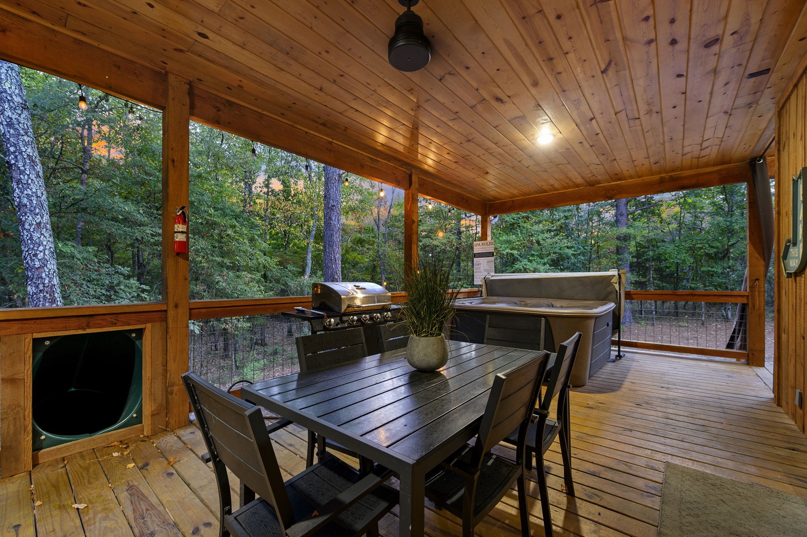 A covered porch sheltered by rich wood beams overlooks peaceful woods dotted with autumn color. The space features a dining table, barbecue, hot tub, and warm lighting—everything for memorable evenings outdoors.