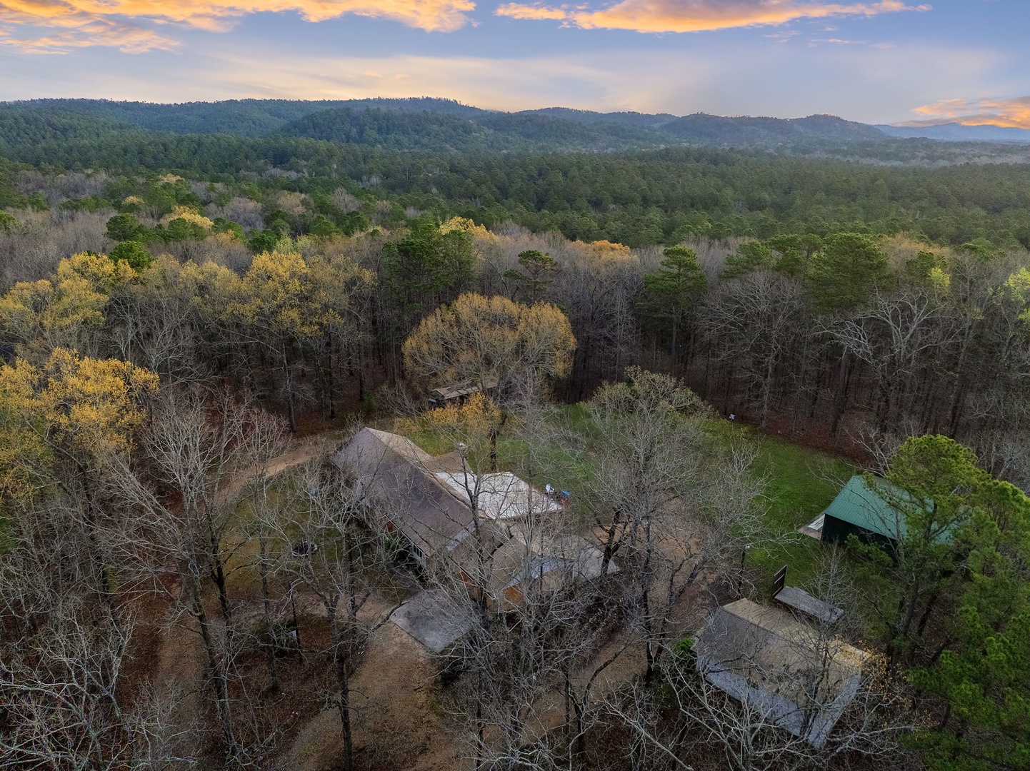 Aerial view captures the property nestled among rolling forested hills beneath a golden sunset sky.
