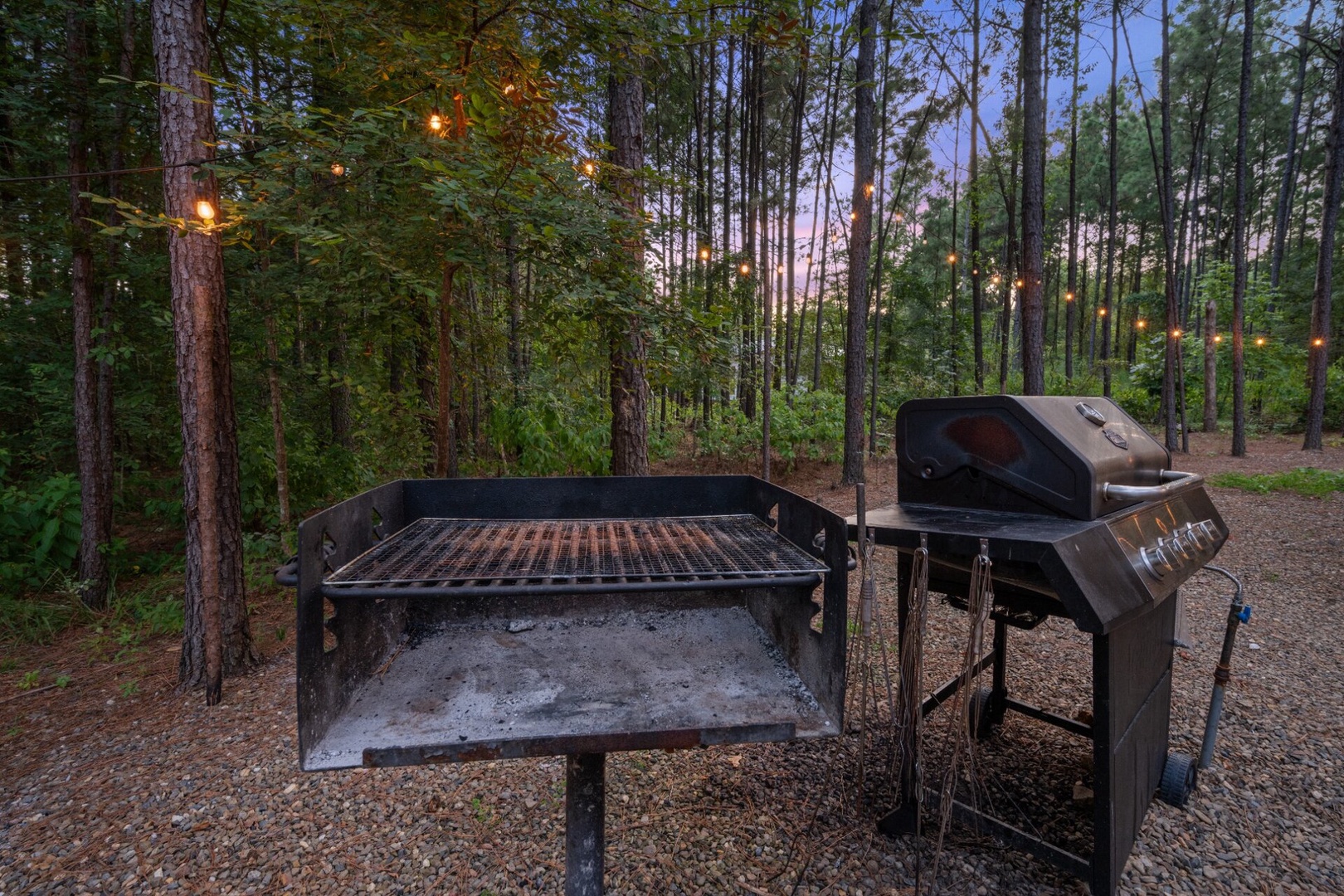 A tranquil wooded area surrounded by towering pines provides the perfect backdrop for outdoor cooking, featuring both a charcoal grill and gas BBQ ready for your cookout under twinkling lights.