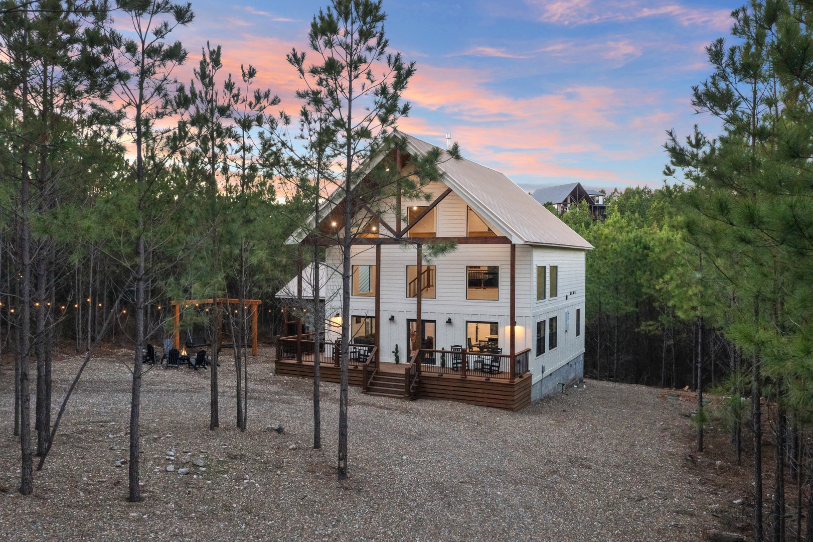 Modern farmhouse retreat nestled among towering pines, featuring wraparound decks and outdoor amenities beneath stunning twilight skies.
