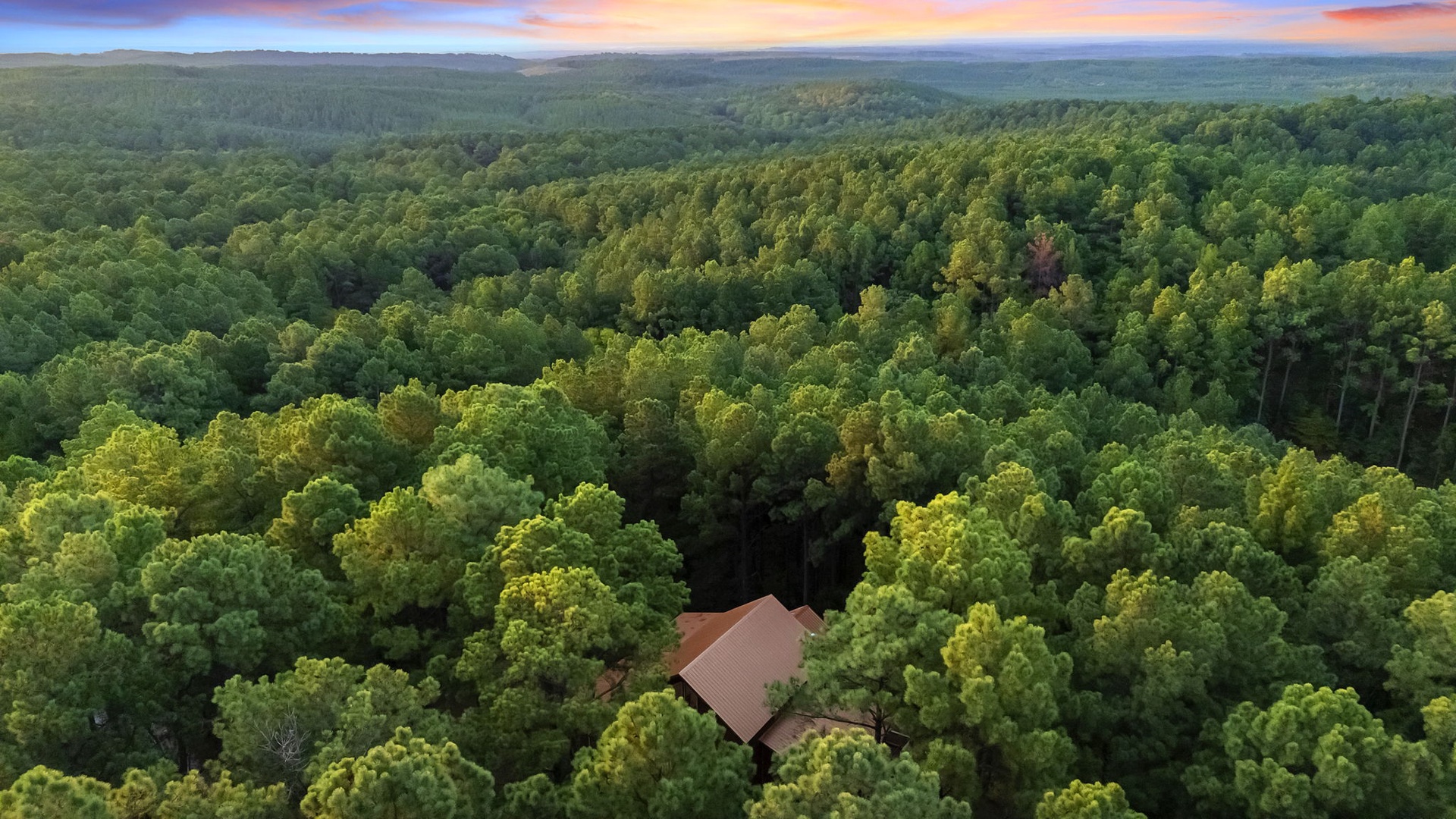 An aerial view captures the property nestled within rolling forested hills, showcasing the secluded natural setting and dramatic sunset skies that frame the surrounding wilderness.