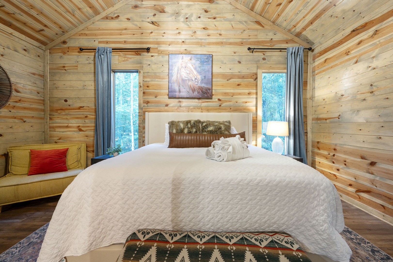 Sink into crisp linens beneath soaring pine ceilings where natural light pours through flanking windows, framing your restful retreat in warm, rustic charm.