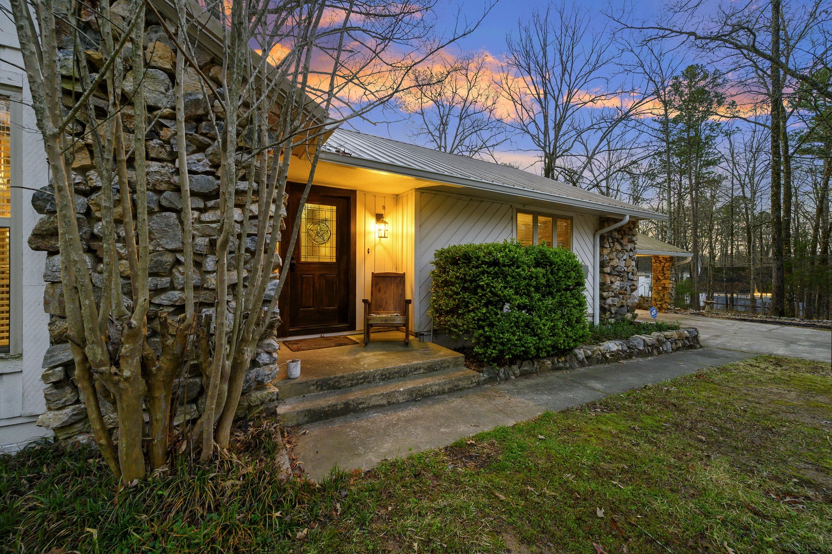 A charming stone and wood cottage surrounded by peaceful forest, featuring warm entrance lighting and natural landscaping.
