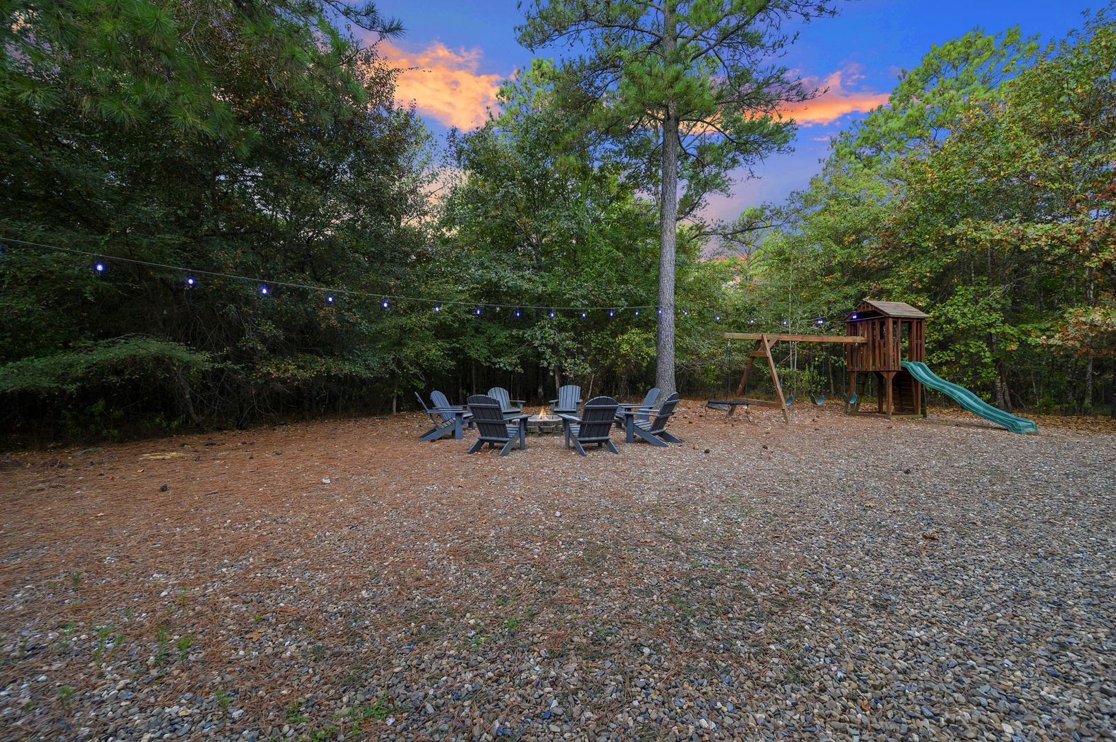 A spacious outdoor area surrounded by lush trees, featuring a gravel gathering spot with Adirondack chairs around a fire pit, strung lights overhead, and a children's playset with swing and slide.