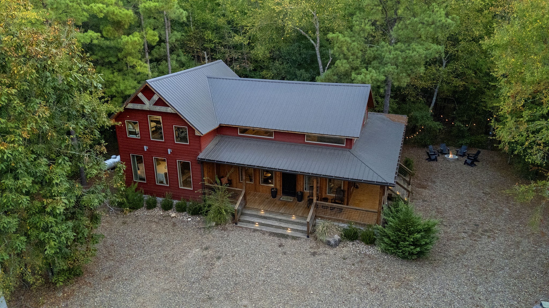 A distinctive red barn-style retreat nestled among lush trees, with gravel parking and a cozy covered porch welcoming guests to this peaceful wooded getaway.