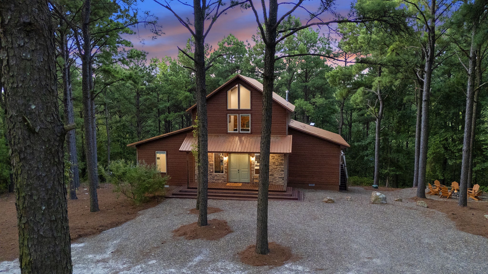 A charming wood-sided cabin nestled among towering pines, glowing warmly as dusk settles over the forest, with a gravel drive and seating area nearby.