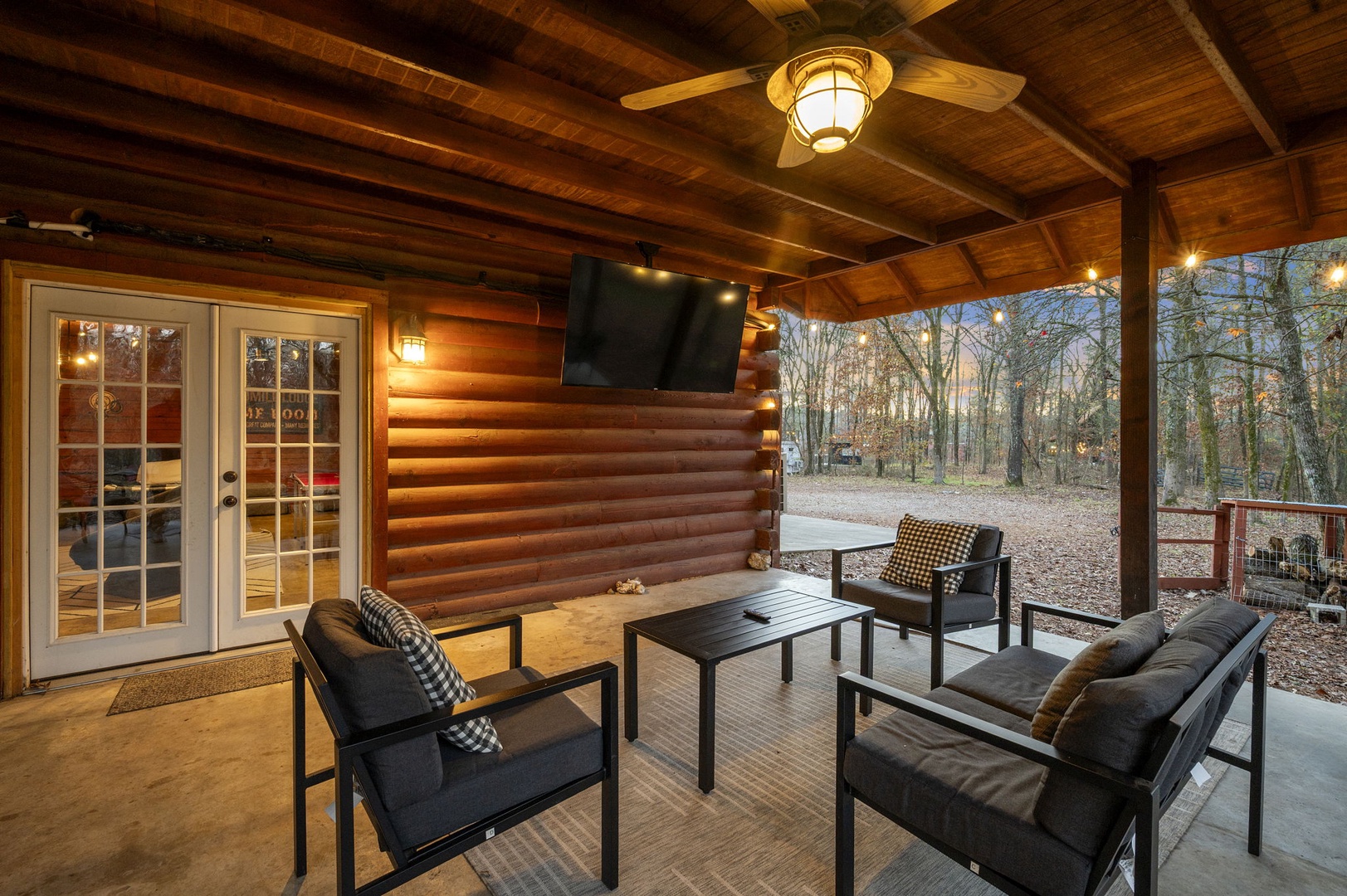Your covered patio invites peaceful evenings with cozy seating, warm wood beams overhead, and serene forest views stretching beyond.