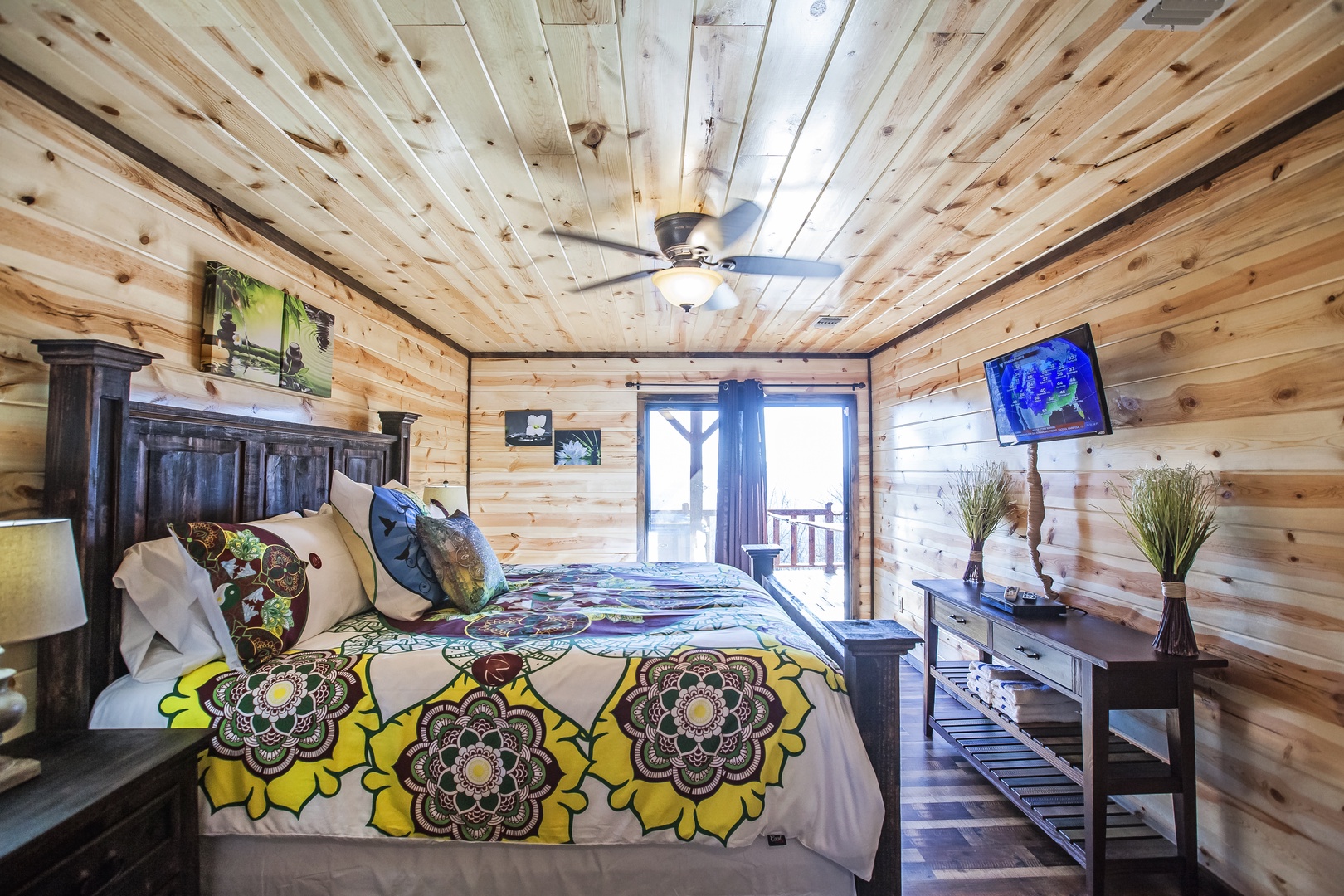 Unwind in your rustic cabin bedroom with warm wood walls, ceiling fan, colorful bedding, and balcony access for peaceful mountain mornings.