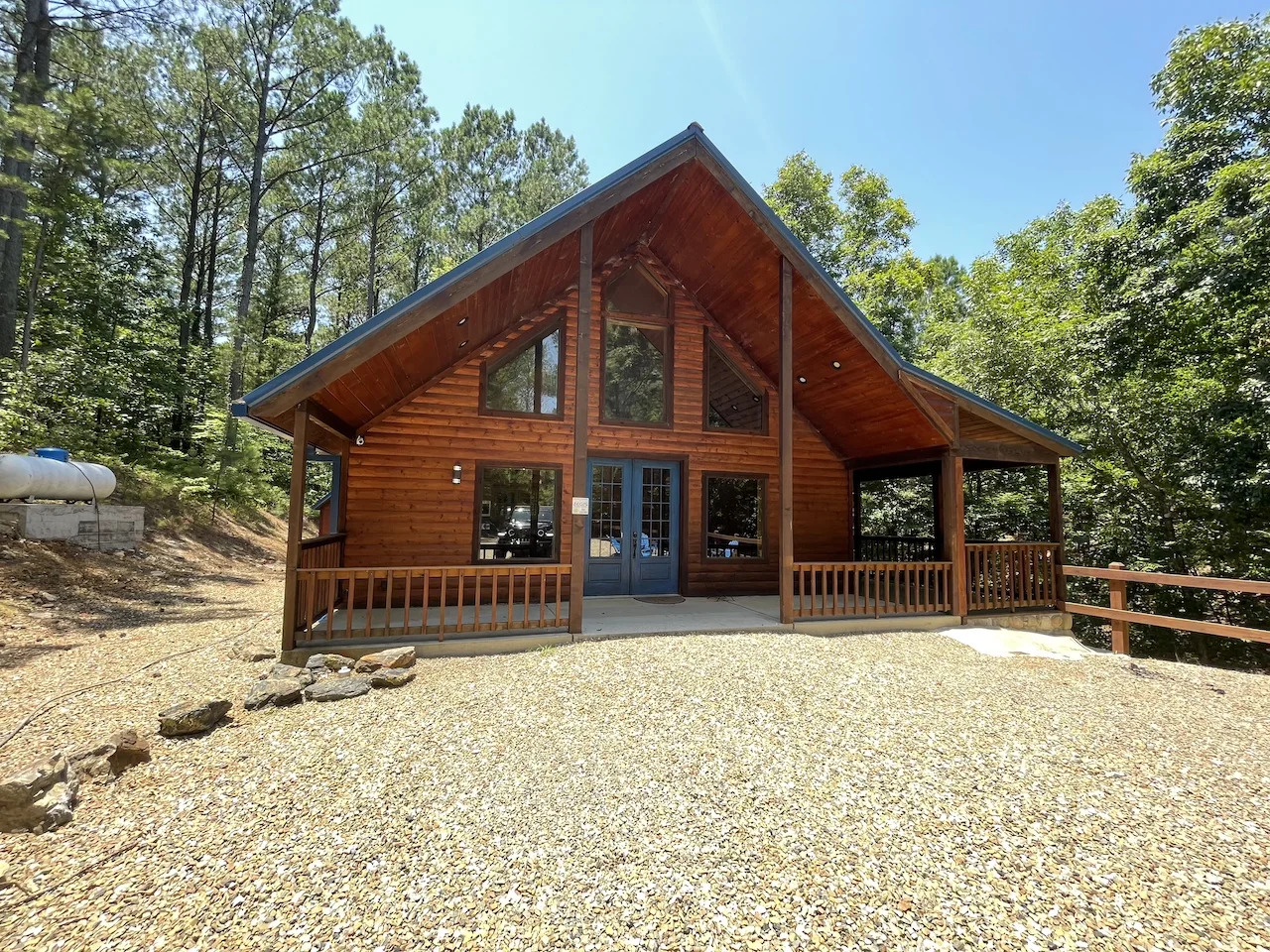 A charming log cabin nestled in the woods features a warm wooden exterior, wraparound deck, and expansive windows that frame the surrounding trees and natural landscape.
