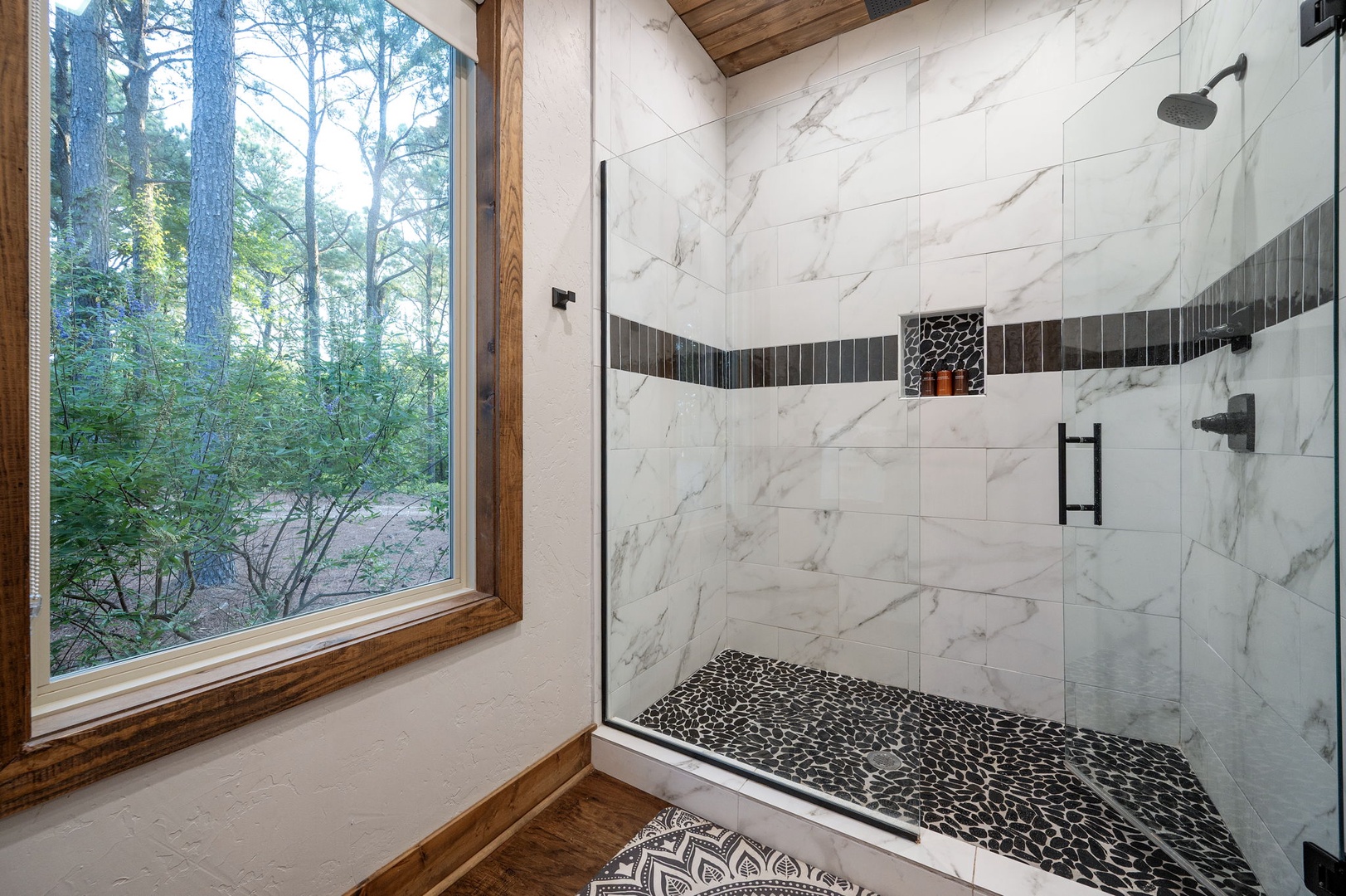 Step into your private spa-like retreat with a gorgeous walk-in shower featuring marble-look tiles and elegant dark accents. Natural light streams through the window, bringing the peaceful forest views right to your morning routine.