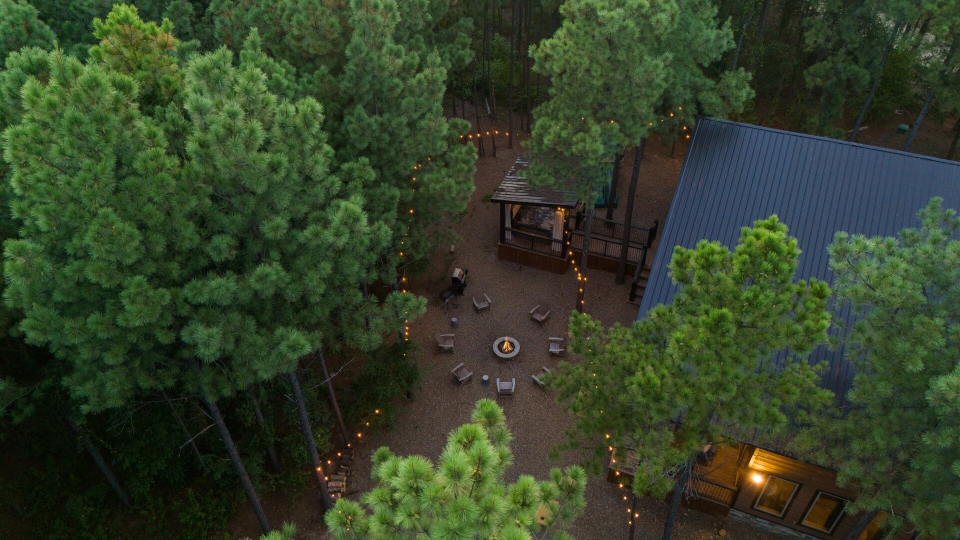 Nestled among tall pines, the property offers a secluded forest retreat with a covered gathering area, fire pit, and enchanting string lights—perfect for peaceful evenings under the trees.