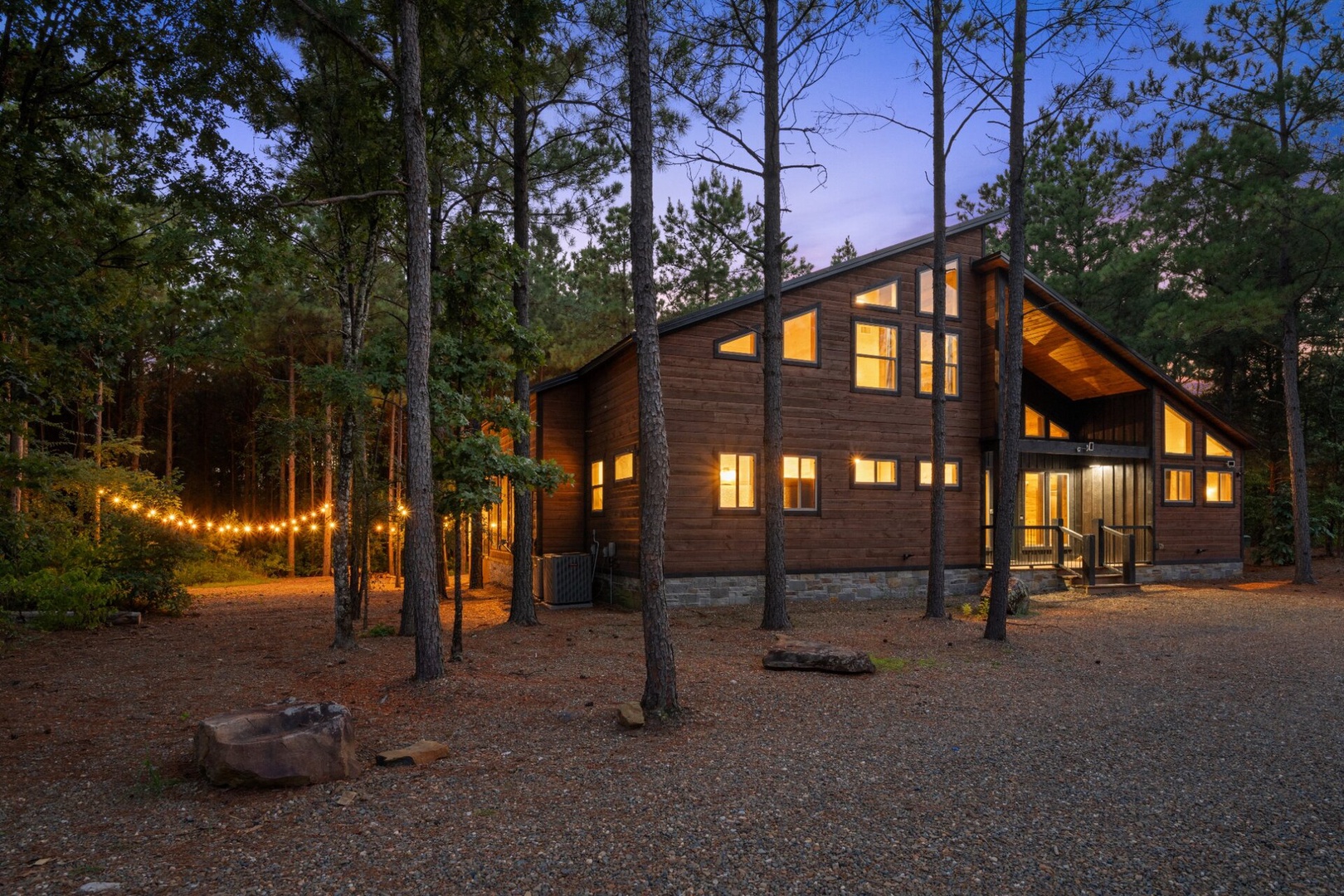 A modern wood-sided cabin glows warmly at twilight, nestled among towering pines with festive string lights welcoming guests to their peaceful forest retreat.