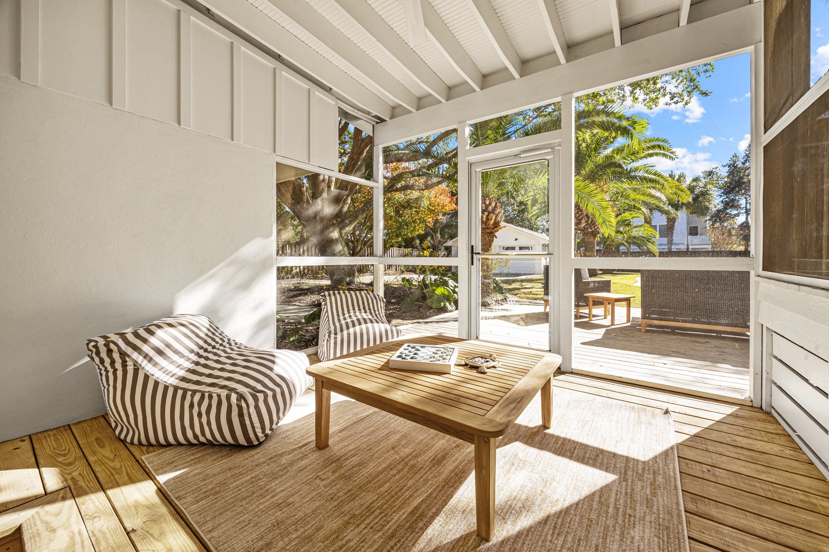 Relax in your sunny enclosed porch with striped cushions and wooden furniture, overlooking tropical palms through floor-to-ceiling windows.