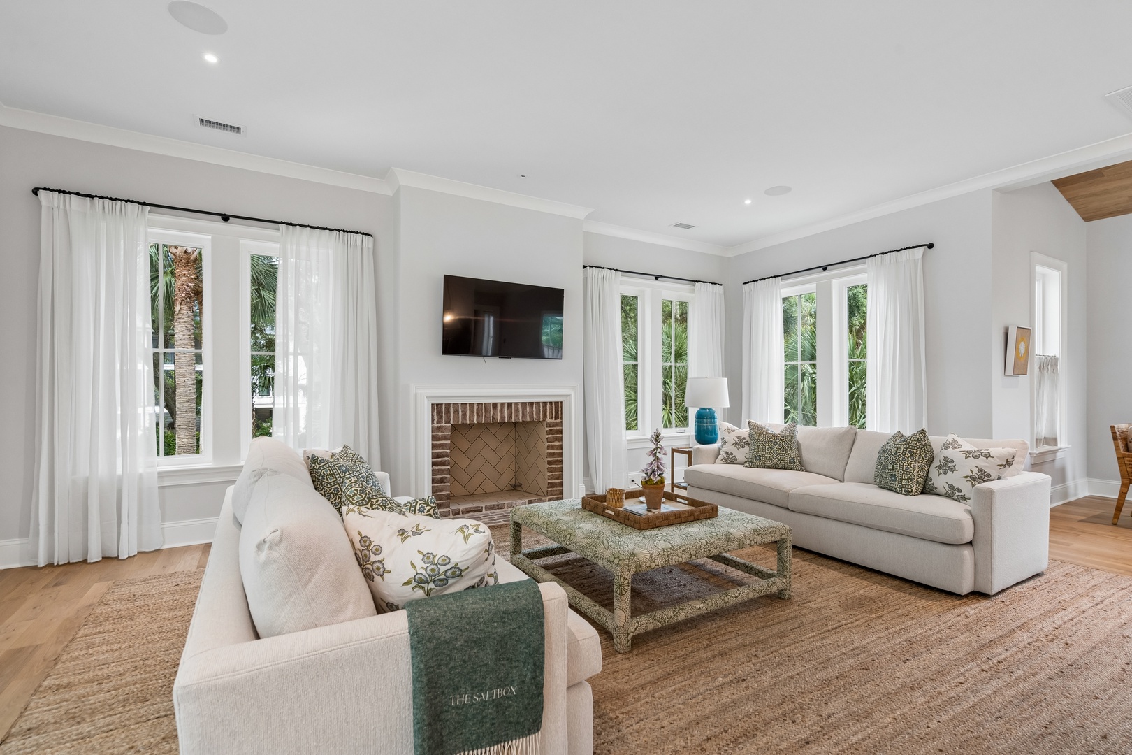 Settle into your bright, airy living room where plush sofas and a warm fireplace invite relaxation. Natural light pours through floor-to-ceiling windows framing lush palm views.