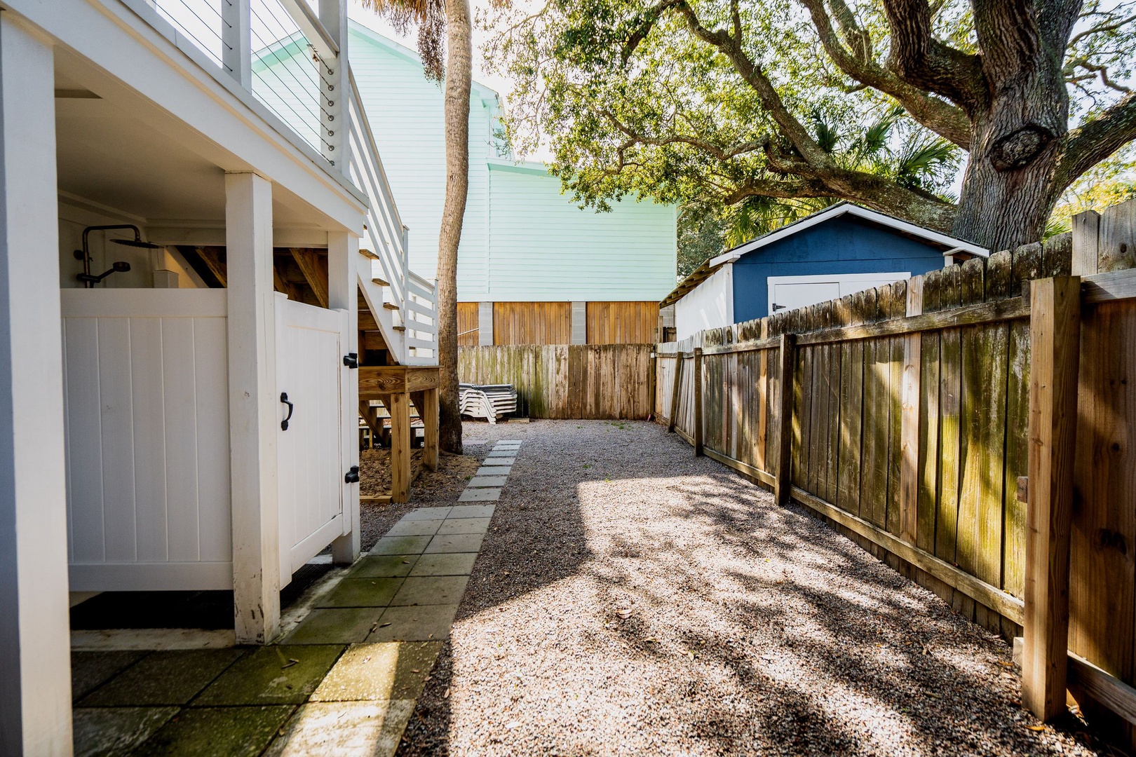 Step into your private backyard sanctuary, where mature trees create natural shade over the gravel courtyard and outdoor shower awaits after beach days.