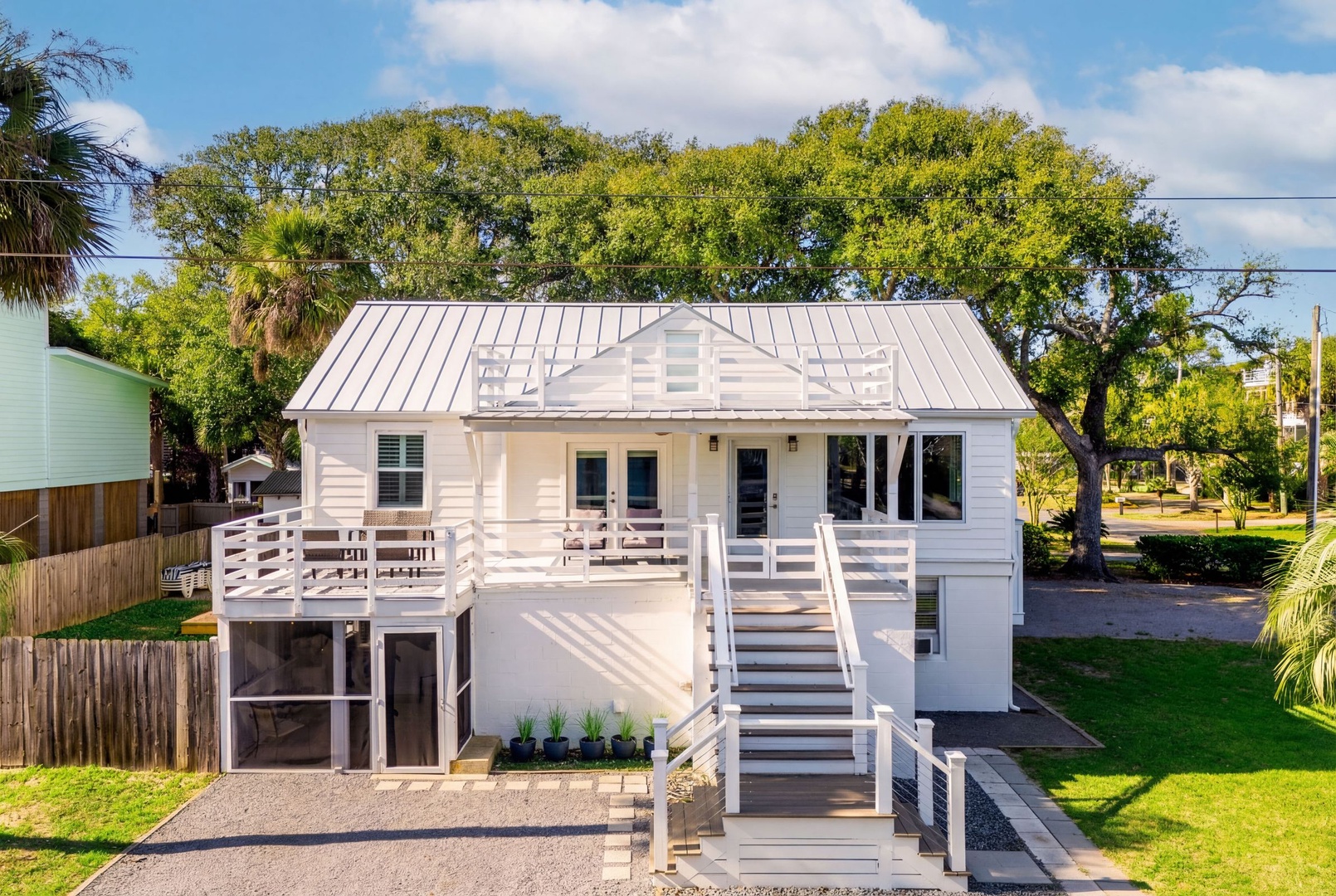 Charming elevated coastal cottage with wraparound deck and metal roof, surrounded by mature oak trees and tropical landscaping.