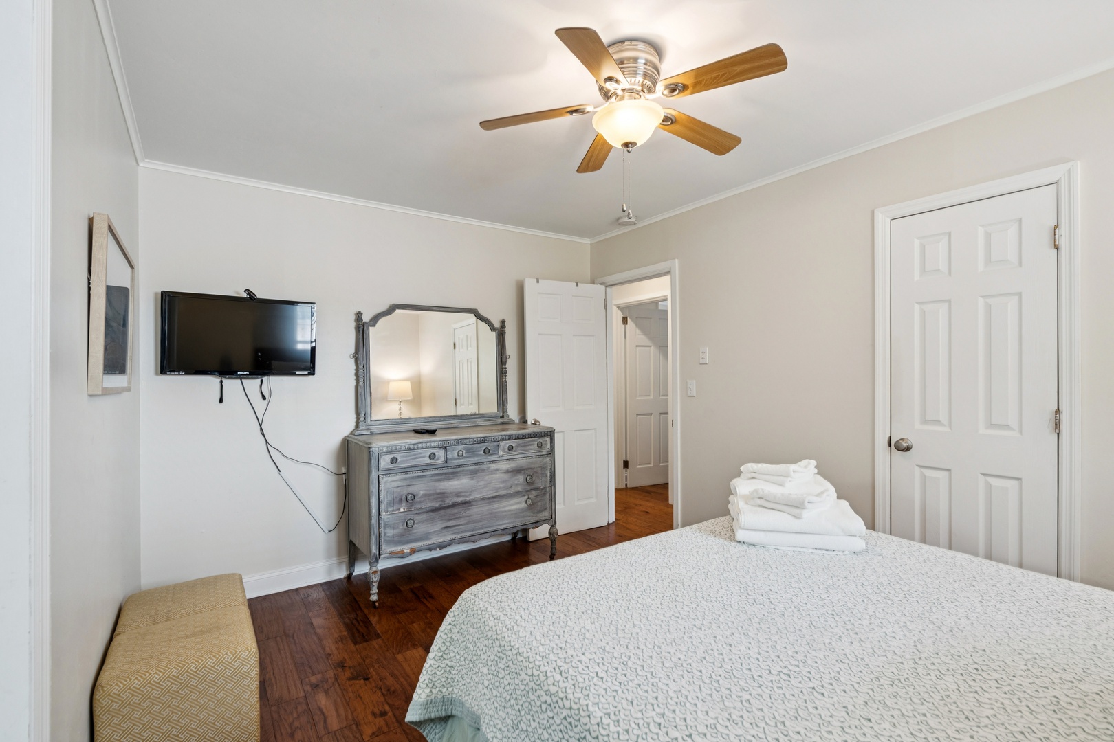 Relax in your cozy bedroom with antique charm, featuring a vintage dresser, wall-mounted TV, and ceiling fan for comfort.