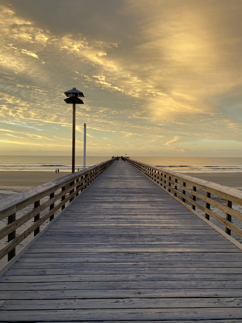 Wooden boardwalk stretches toward golden sunset over calm waters, creating a serene waterfront setting.
