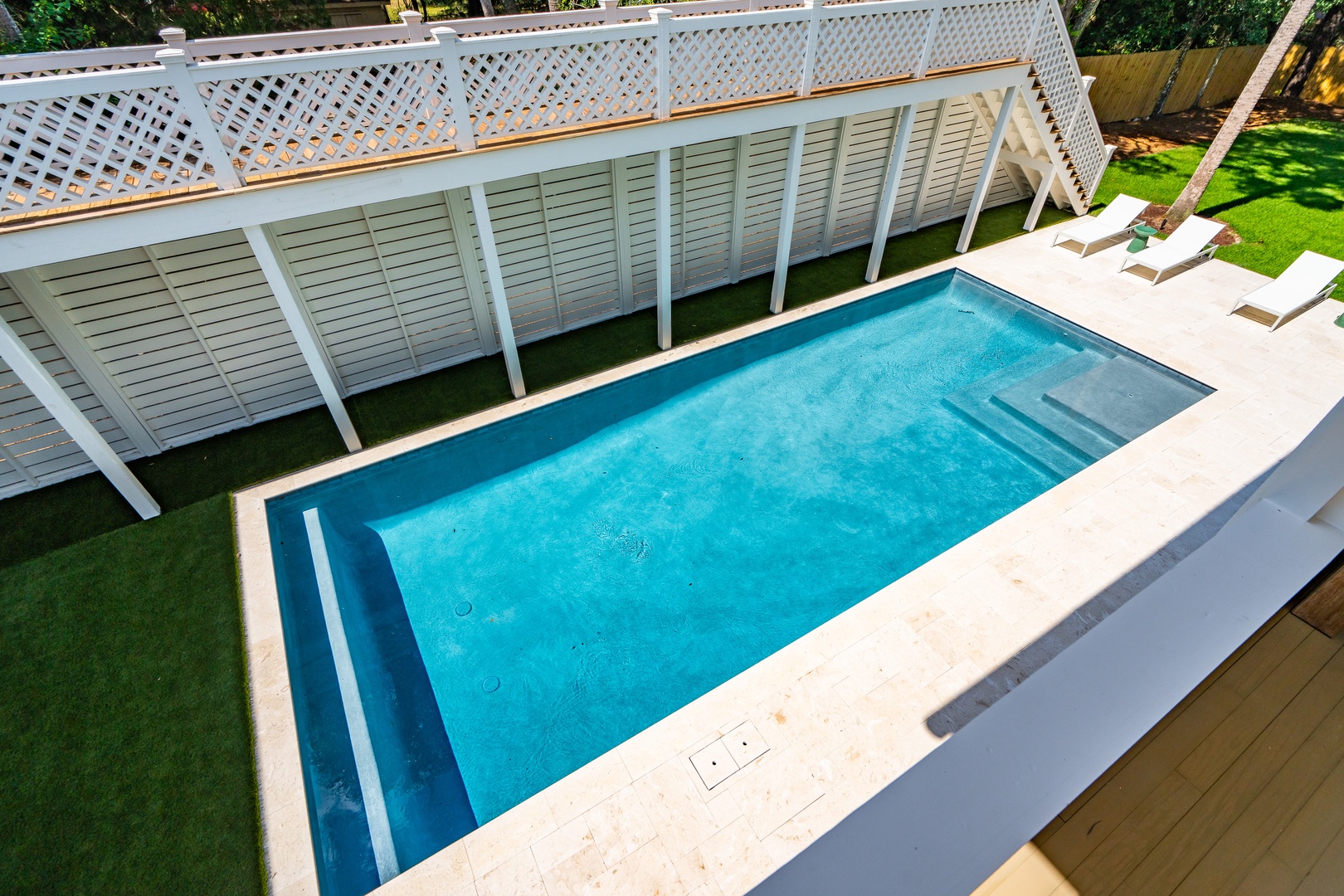 An elevated view of your private swimming pool, framed by pristine white lattice fencing and lush green lawns, with inviting sunbeds awaiting your arrival.
