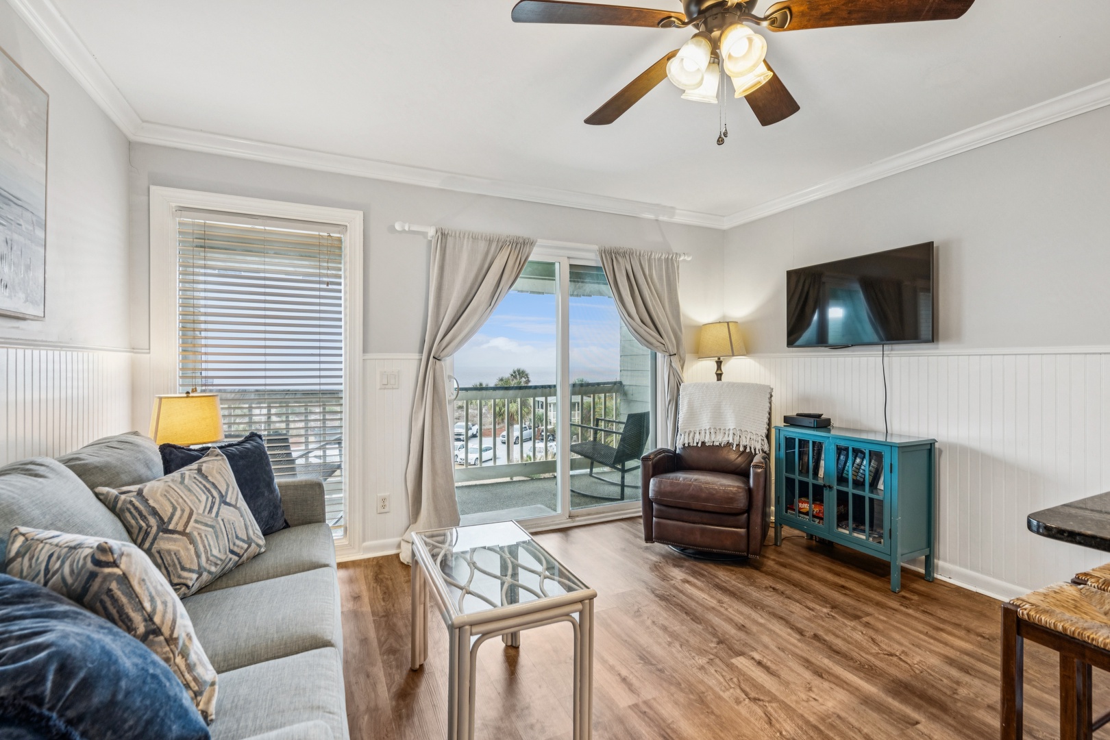 Relax in your bright living room with comfortable seating, ceiling fan, and private balcony access overlooking peaceful surroundings.